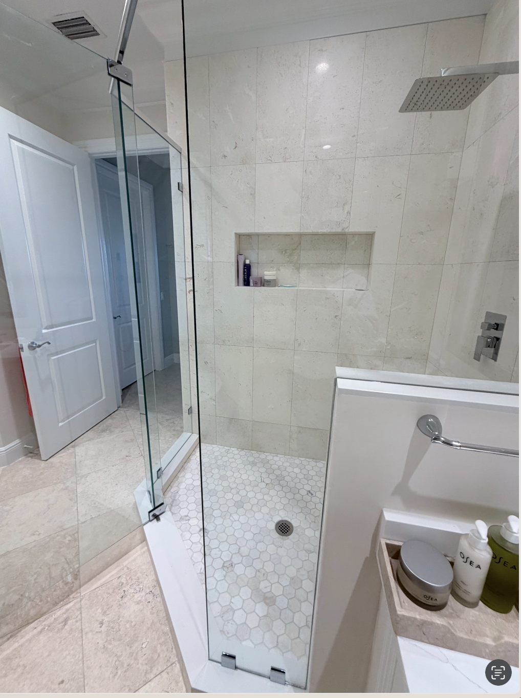 Walk-in shower with beige tile walls and hexagonal tile floor, enclosed by a glass door, with a built-in shelf containing toiletries, at the bathroom entrance.
