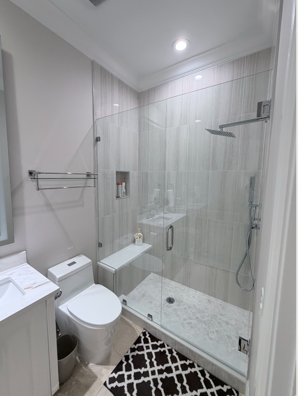A modern bathroom with a glass-enclosed shower, a white toilet, a black and white patterned rug, and a small cabinet with toiletries inside.