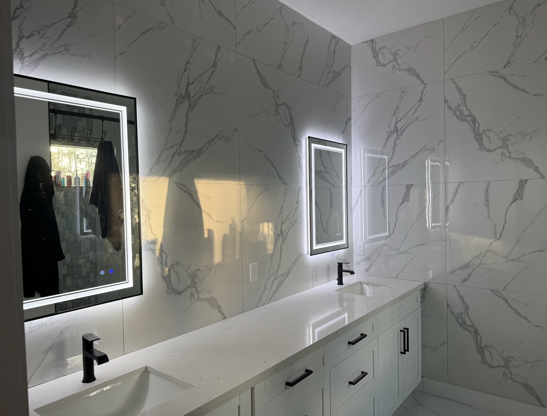 Modern bathroom with white marble walls and countertop, two rectangular mirrored vanities with LED lighting, black faucets, and white cabinetry.