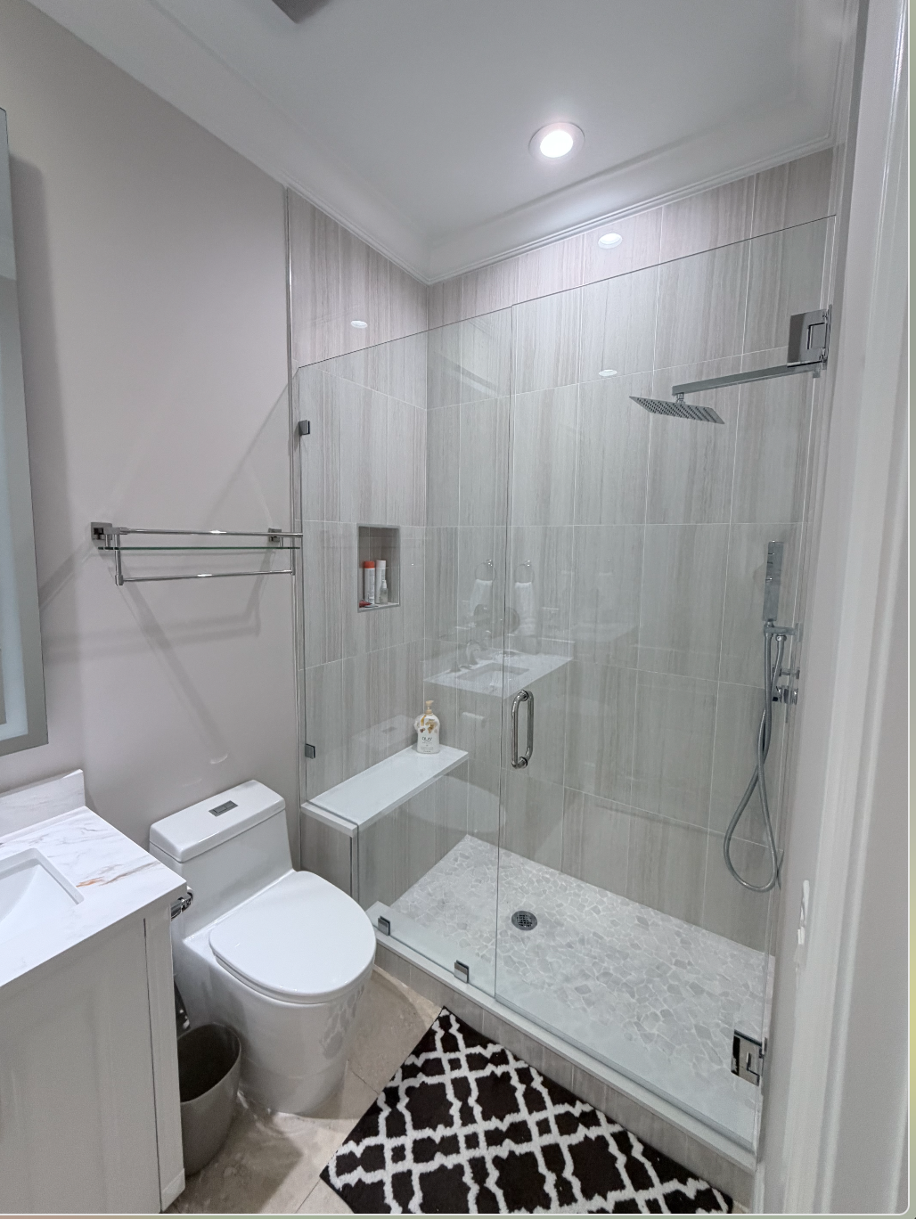 Modern bathroom with enclosed glass shower, toilet, small white cabinet, and patterned rug on beige tile floor.