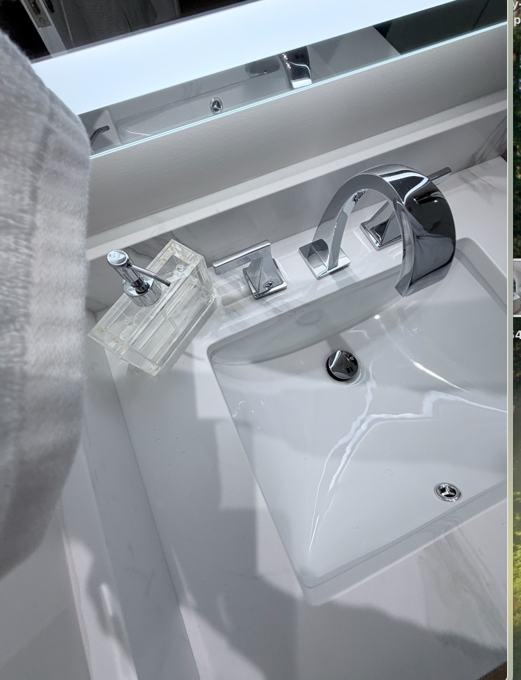 Close-up view of a modern white bathroom sink with a shiny chrome faucet and accessories, including a soap dispenser and mirror.