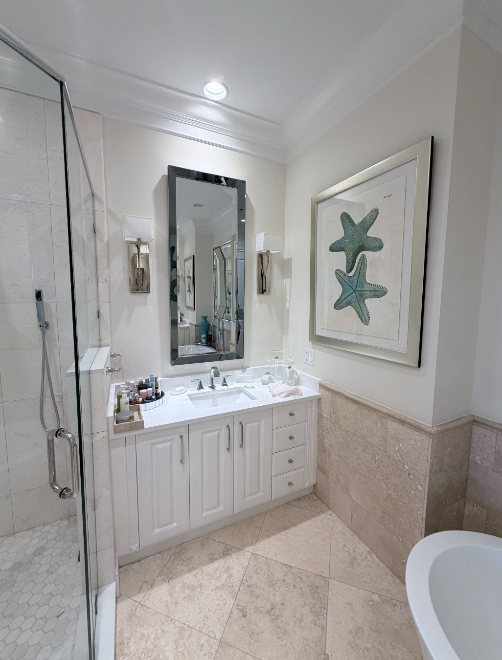 A bathroom with a shower, a white vanity with a mirror, wall sconces, and sea star artwork.