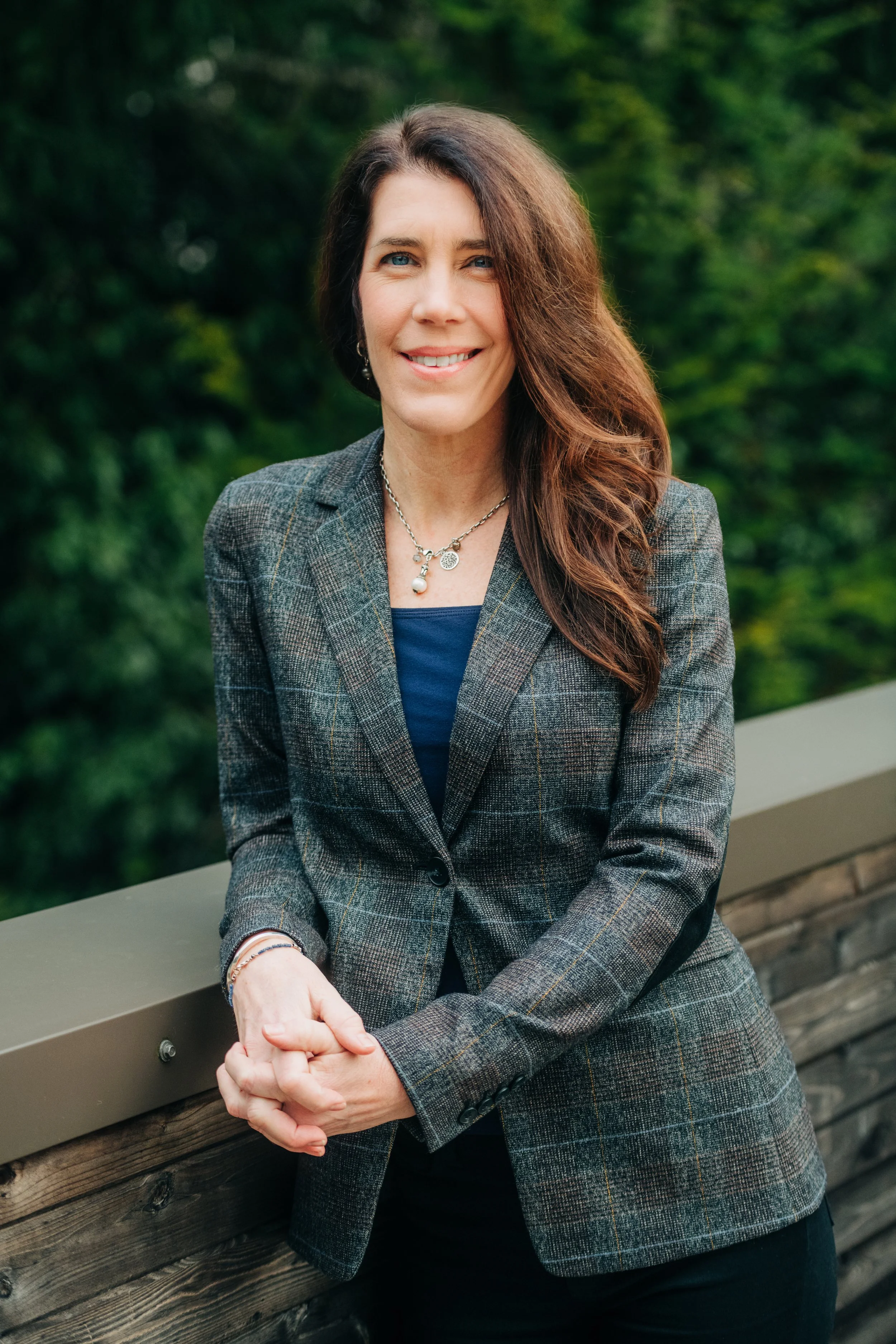 A woman with long brown hair, wearing a plaid blazer over a navy shirt, standing outdoors by a wooden railing with green foliage in the background.