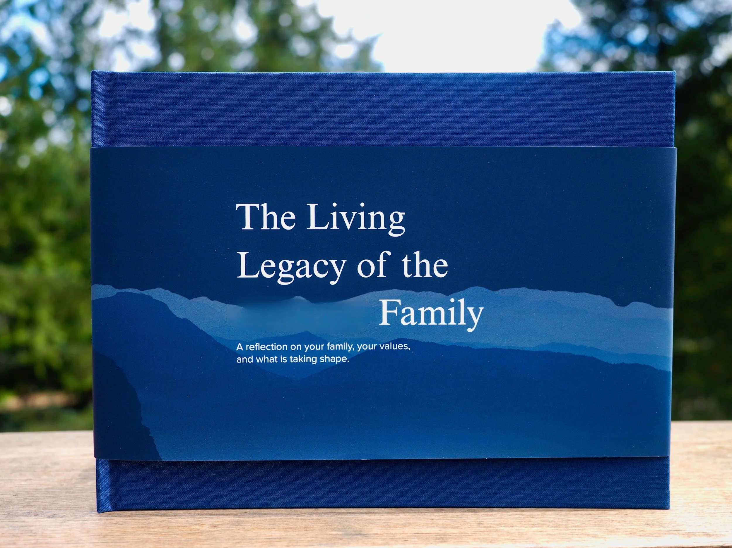 Blue book titled "The Living Legacy of the Family" with subtitle "A reflection on your family, your values, and what is taking shape" in an outdoor setting with trees in the background.