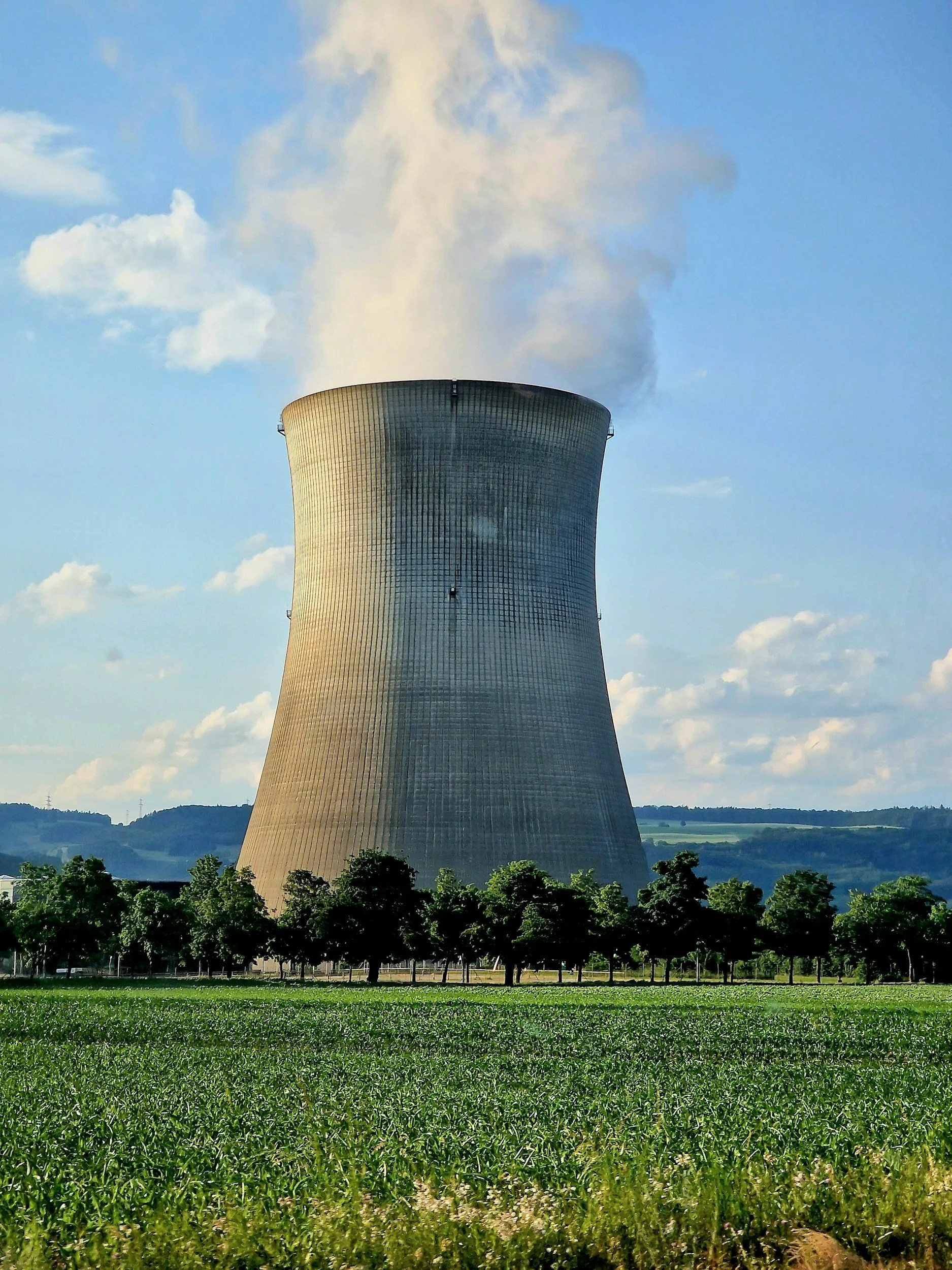 A nuclear power plant cooling tower emitting steam, set against a partly cloudy sky and surrounded by green trees and fields.