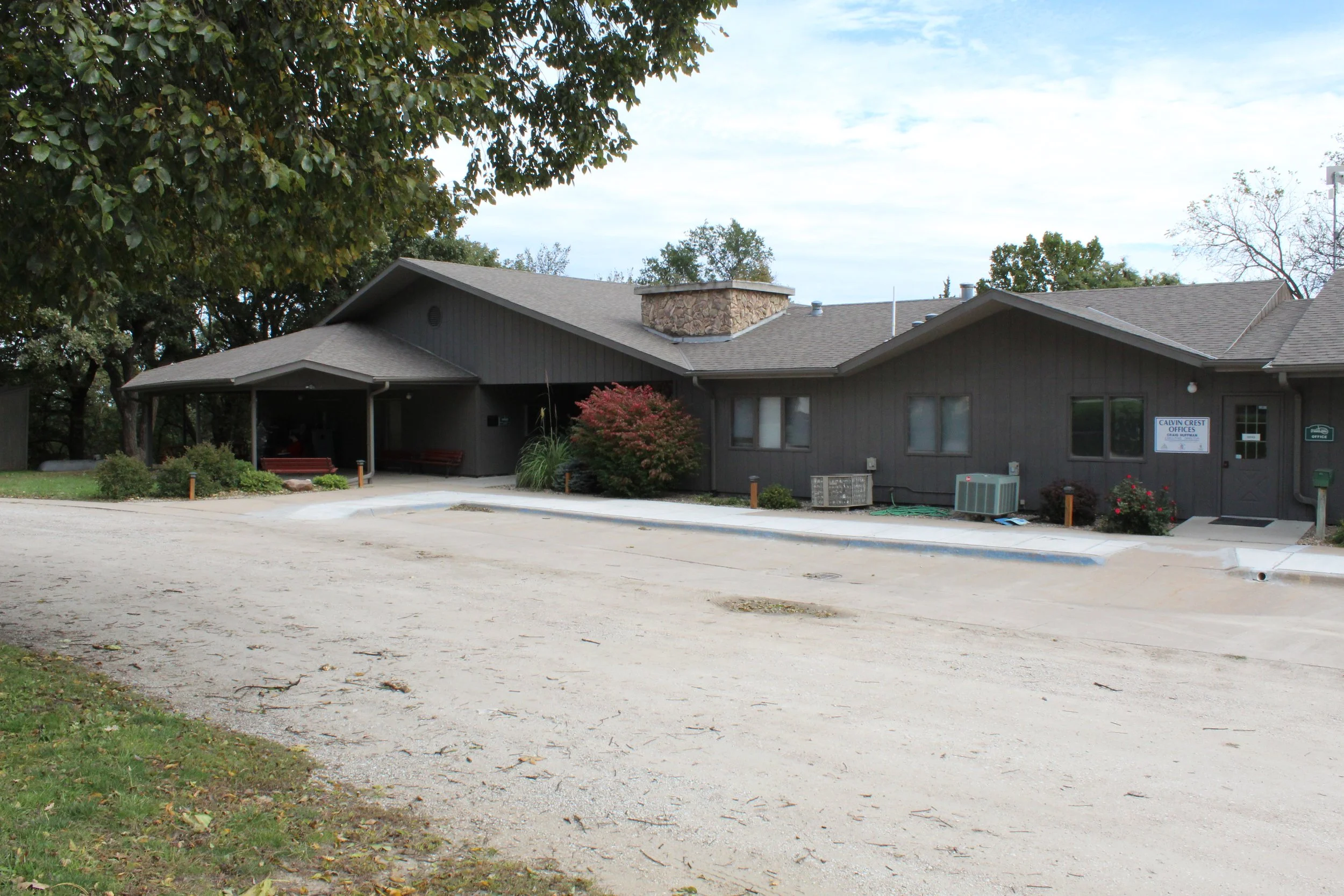 A single-story building with a dark gray exterior, multiple windows, and a small porch area with benches, buildings surrounded by trees, a gravel parking lot in front, and a sign indicating this is Calvin Crest.