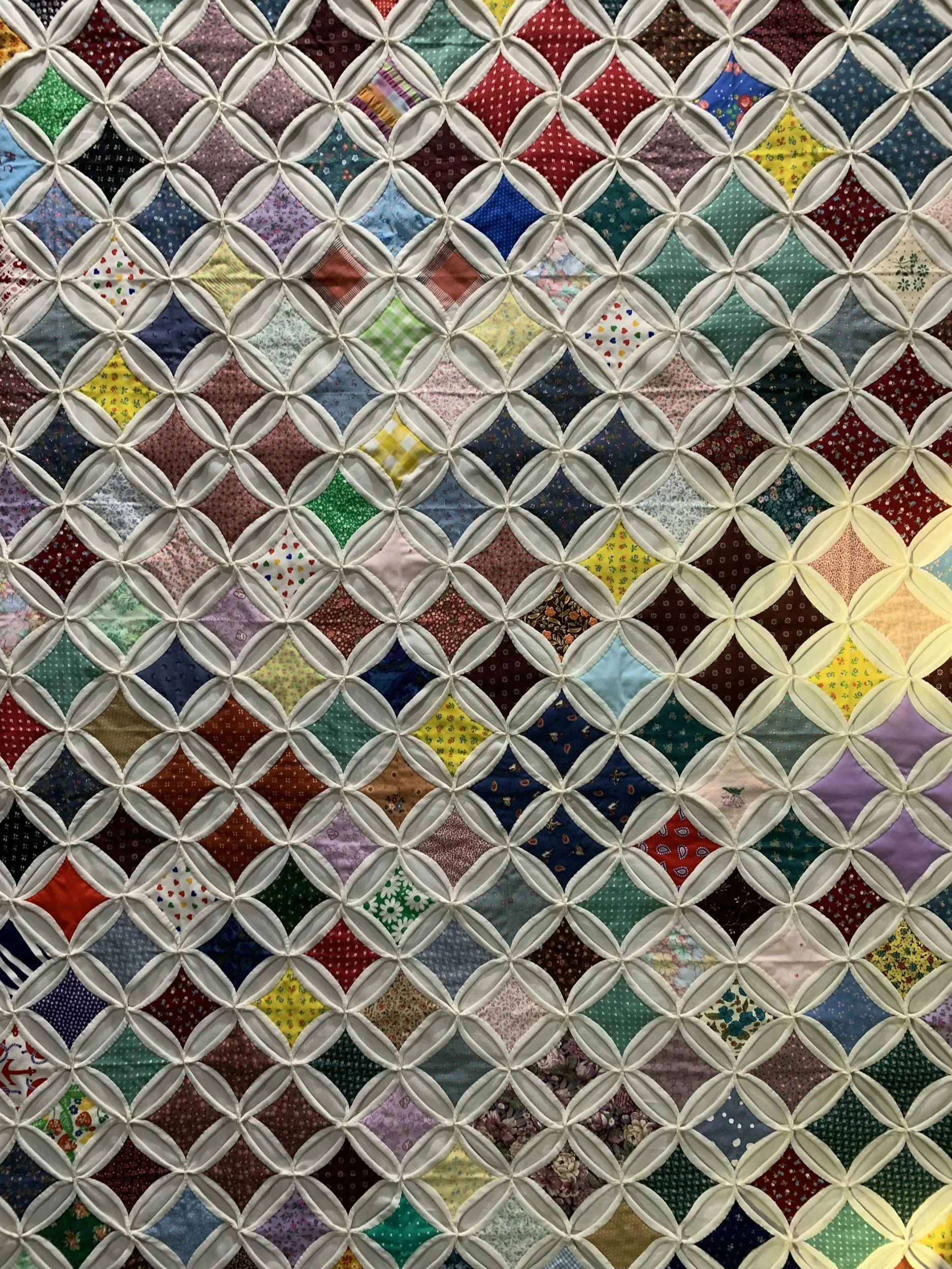 Close-up of a quilt made of small, colorful fabric squares arranged in a diamond pattern with a white lattice quilting overlay.
