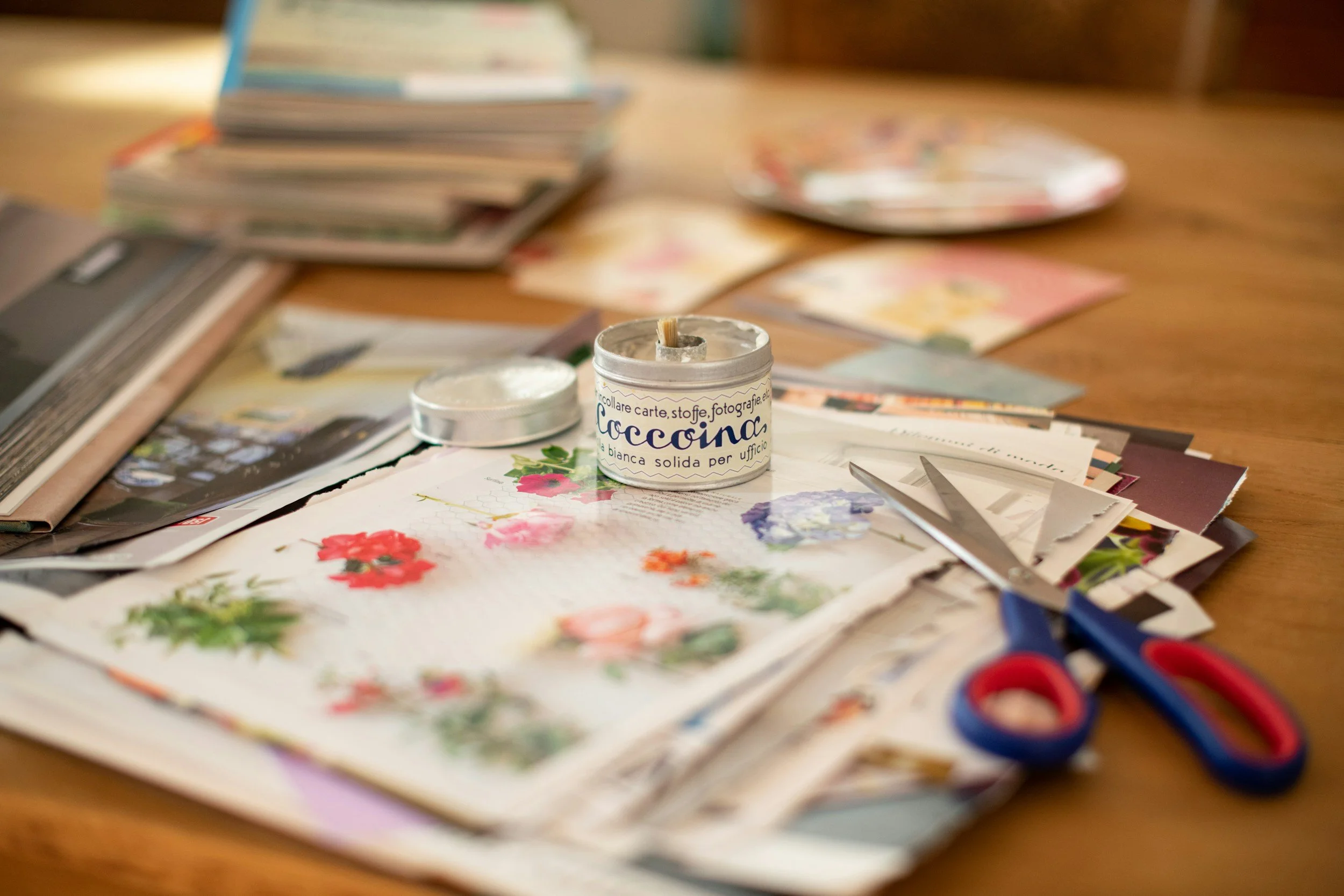 A craft table with floral paper, scissors, and a small candle labeled "Laccina" among scattered papers and photos.