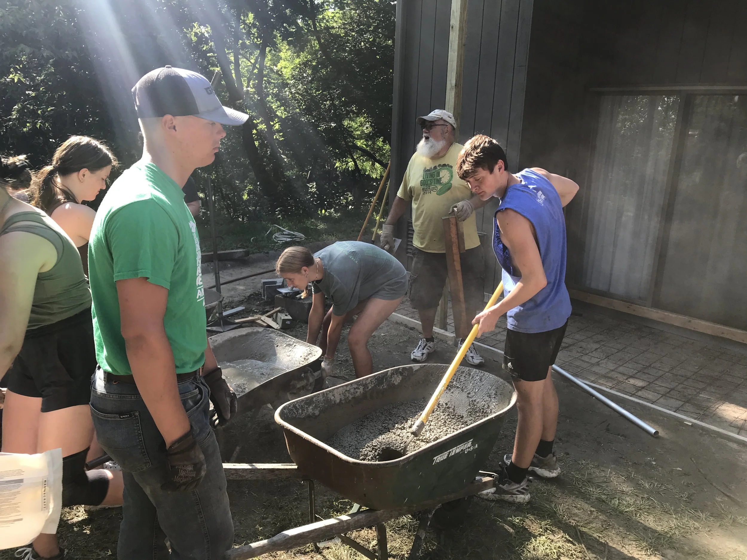 Group of people working together outdoors, mixing cement in wheelbarrows during daylight.