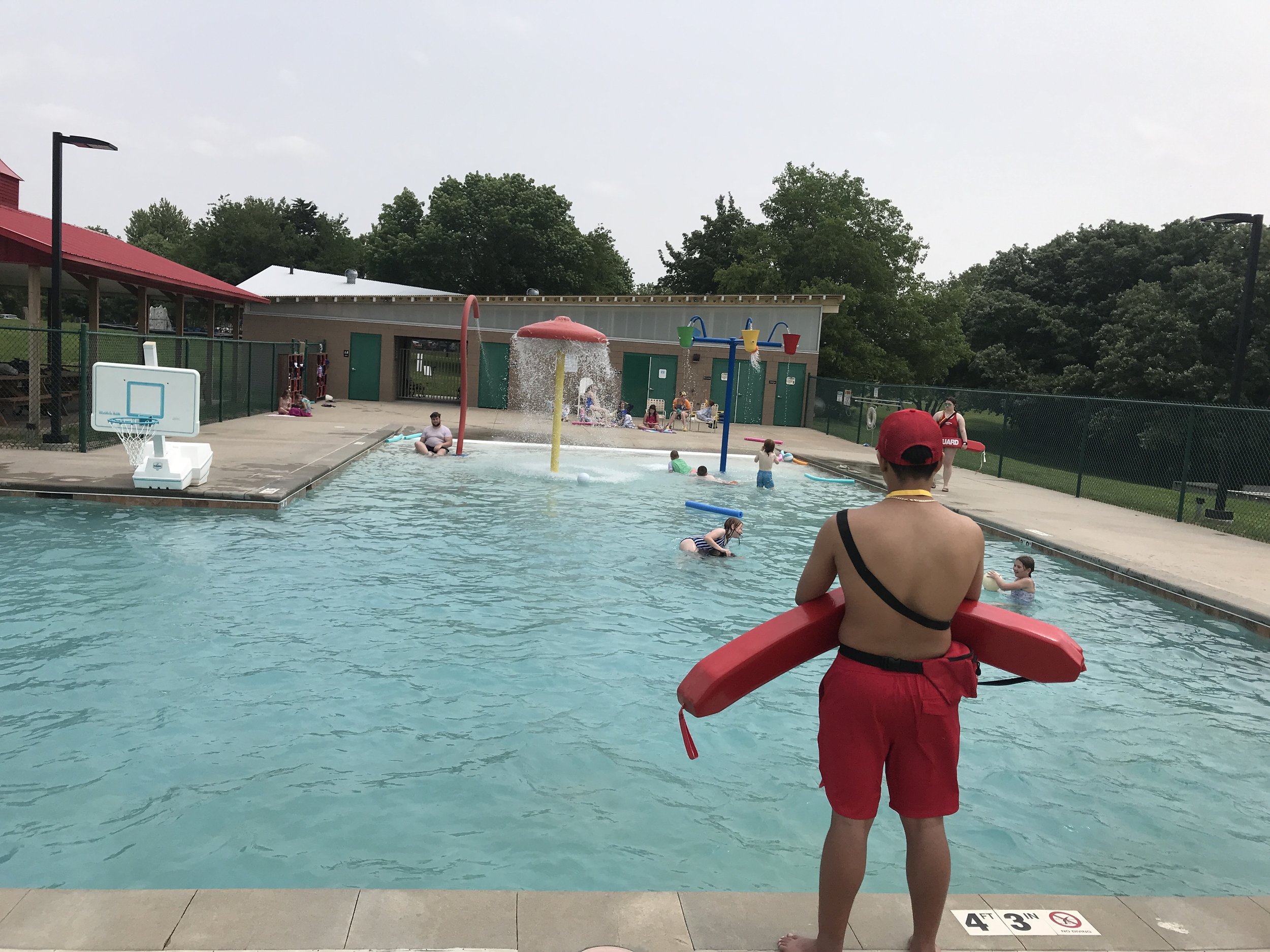 A lifeguard in red shorts and a red hat watches over children swimming and playing in an outdoor pool, with water playground equipment and a fenced grassy area in the background.