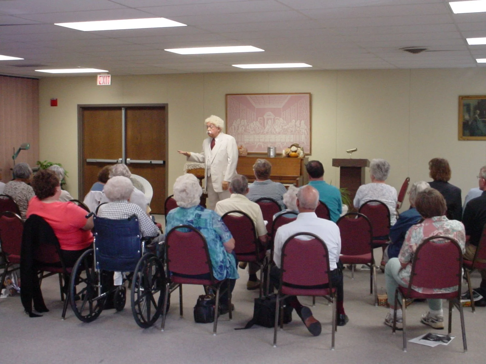 A man dressed as Mark Twain giving a lecture to a group of elderly people, some in wheelchairs, in a community center with framed artwork on the walls.