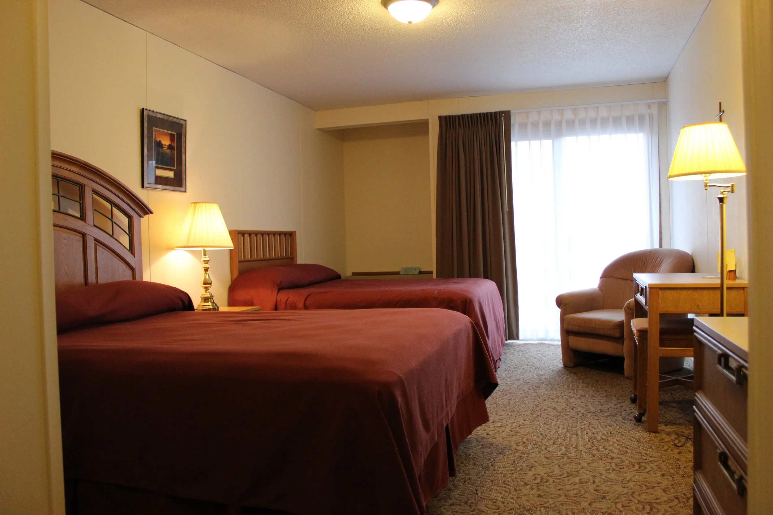 Interior of a hotel room with two beds, lamps, a chair, and a sliding glass door with curtains.