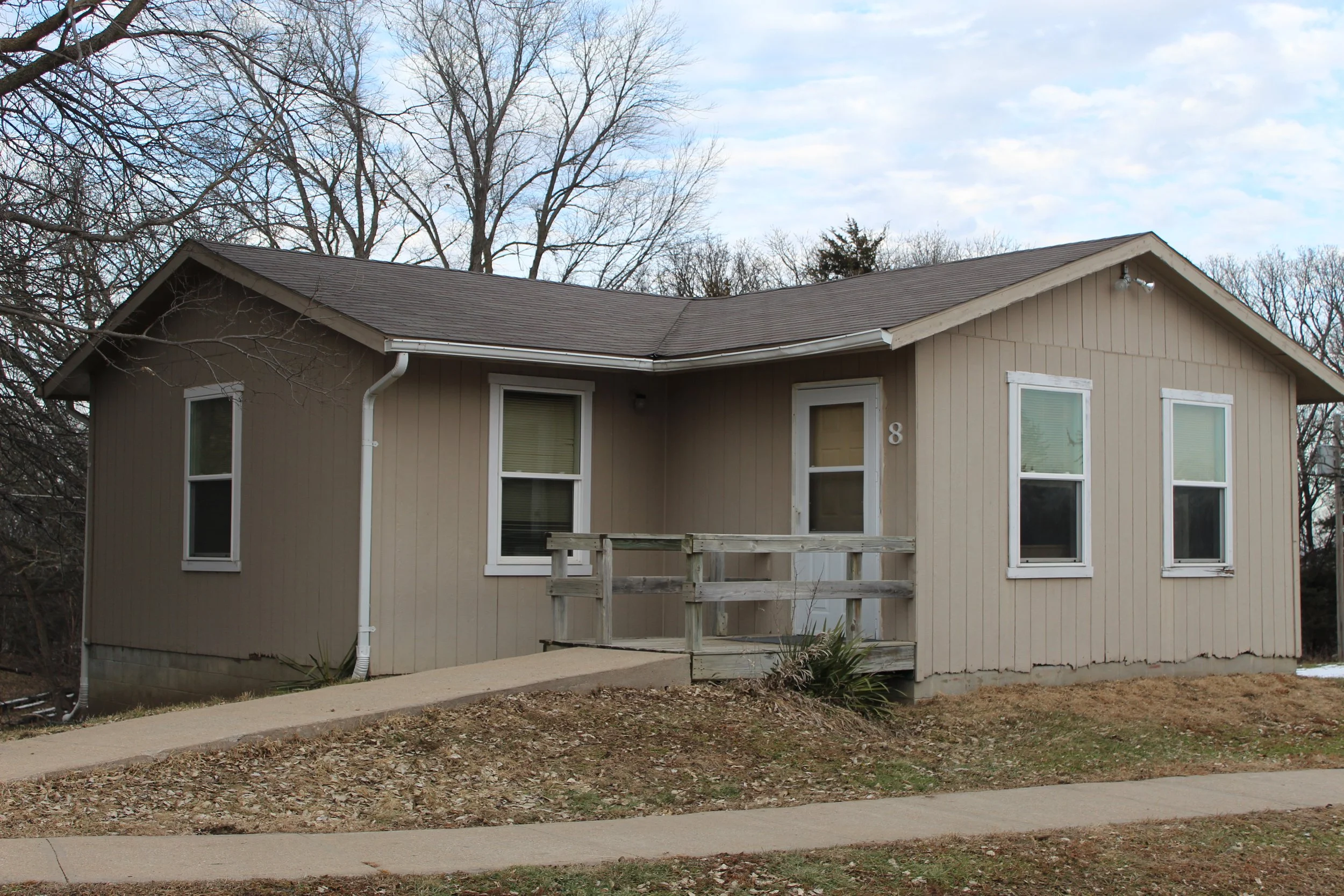 A beige one-story cabin with white trim and a small front porch with wooden railing. The house has four windows and a small ramp leading up to the front door. The house number is 8, and it is surrounded by leafless trees and a partly cloudy sky.