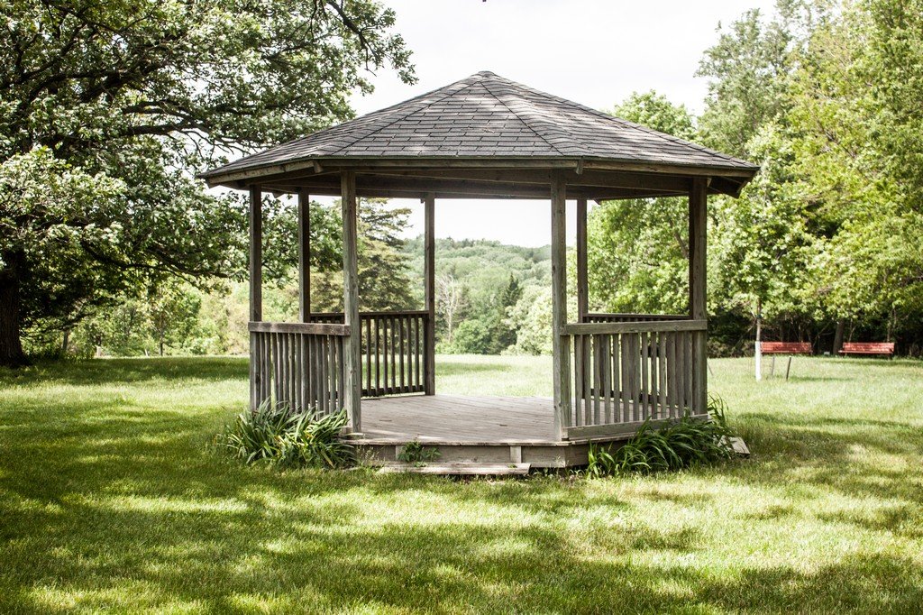 Wooden gazebo in a park surrounded by trees and green grass.