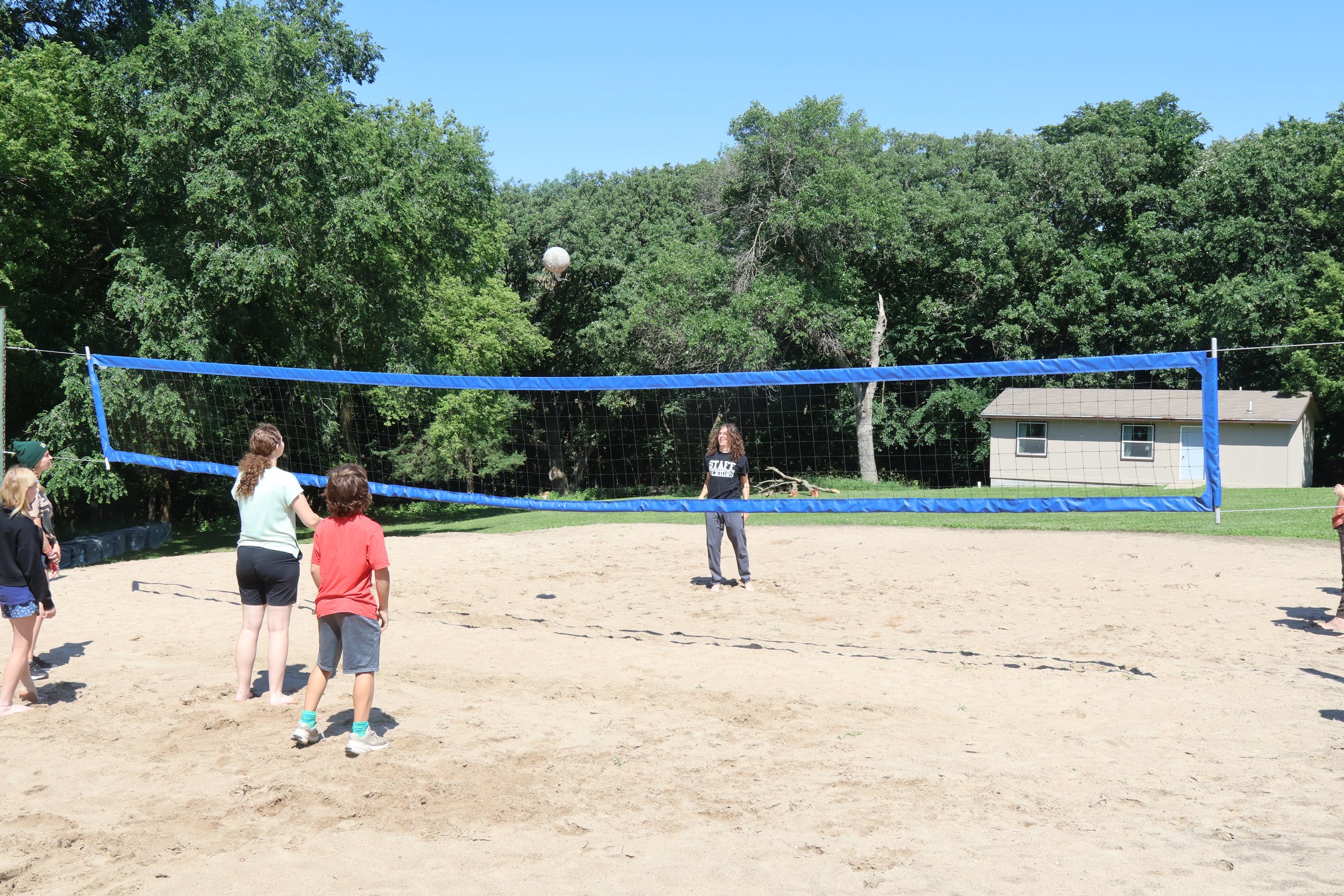 People playing volleyball on a sand court outdoors, with trees and a cabin in the background.