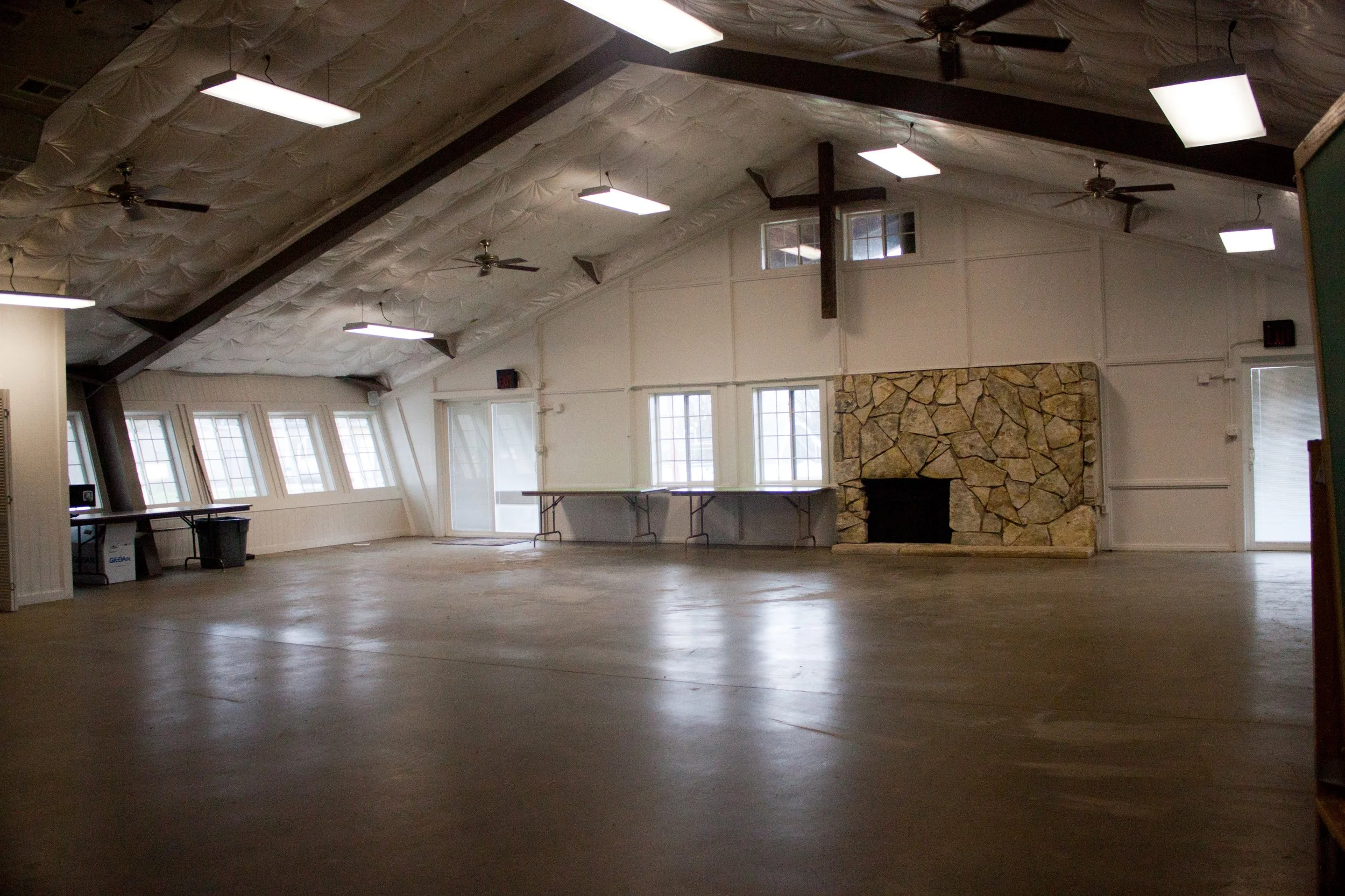 Large, empty gathering hall with a sloped ceiling, multiple windows, ceiling fans, and a stone fireplace.