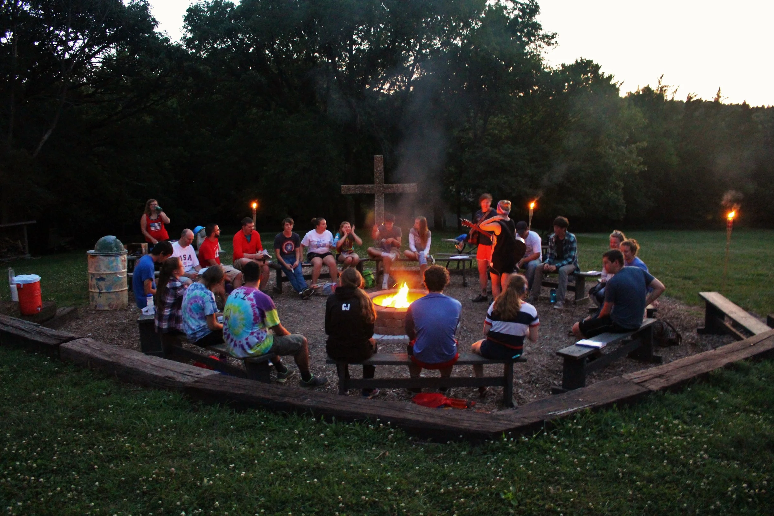 A group of people sitting around a campfire in a circle outdoors during the evening. Some are playing musical instruments, and tiki torches line the background. There is a large cross in the background.