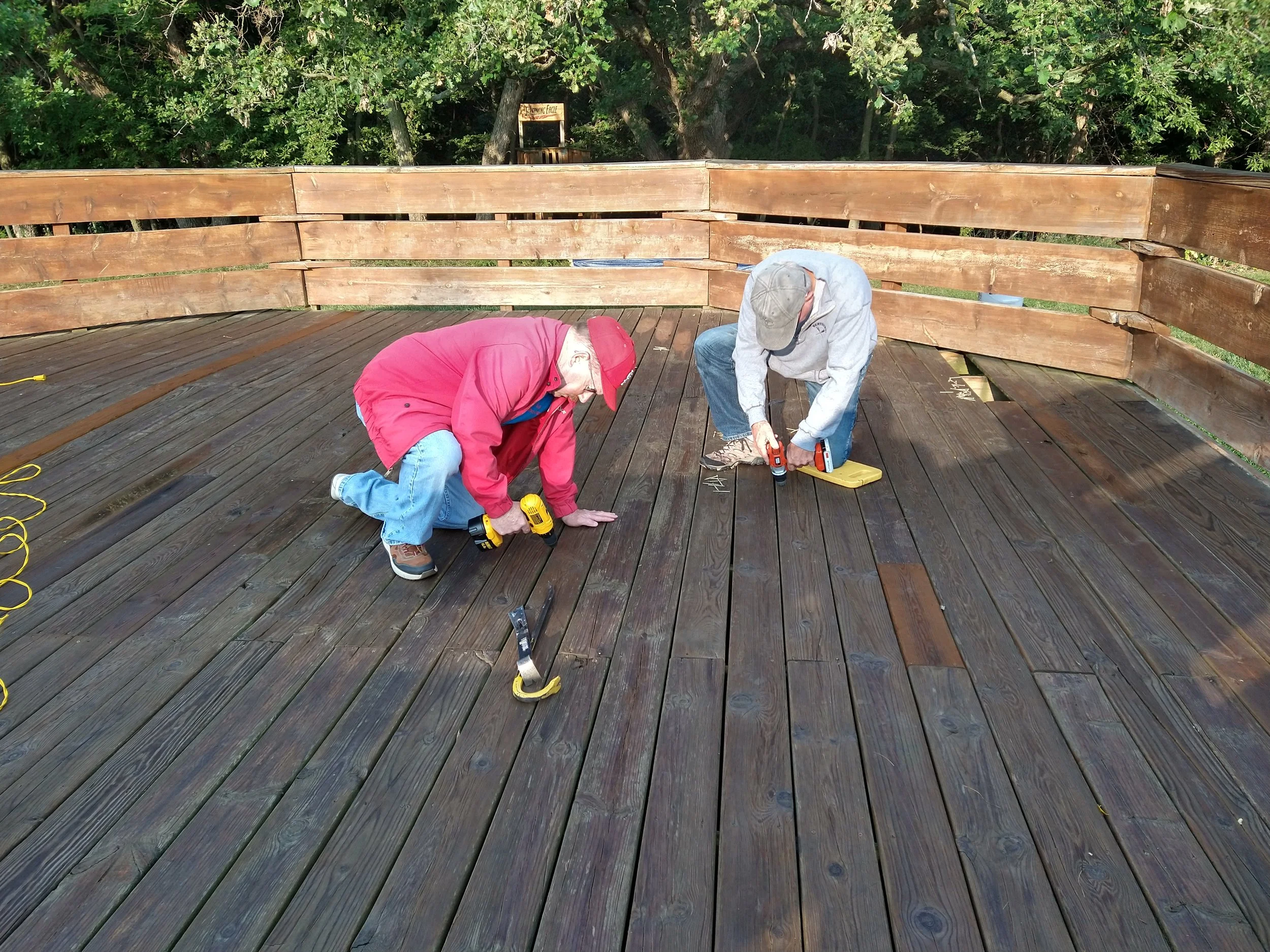 Two men working on installing or repairing a wooden deck outdoors, using power tools, with focus on the deck surface and surrounding greenery.