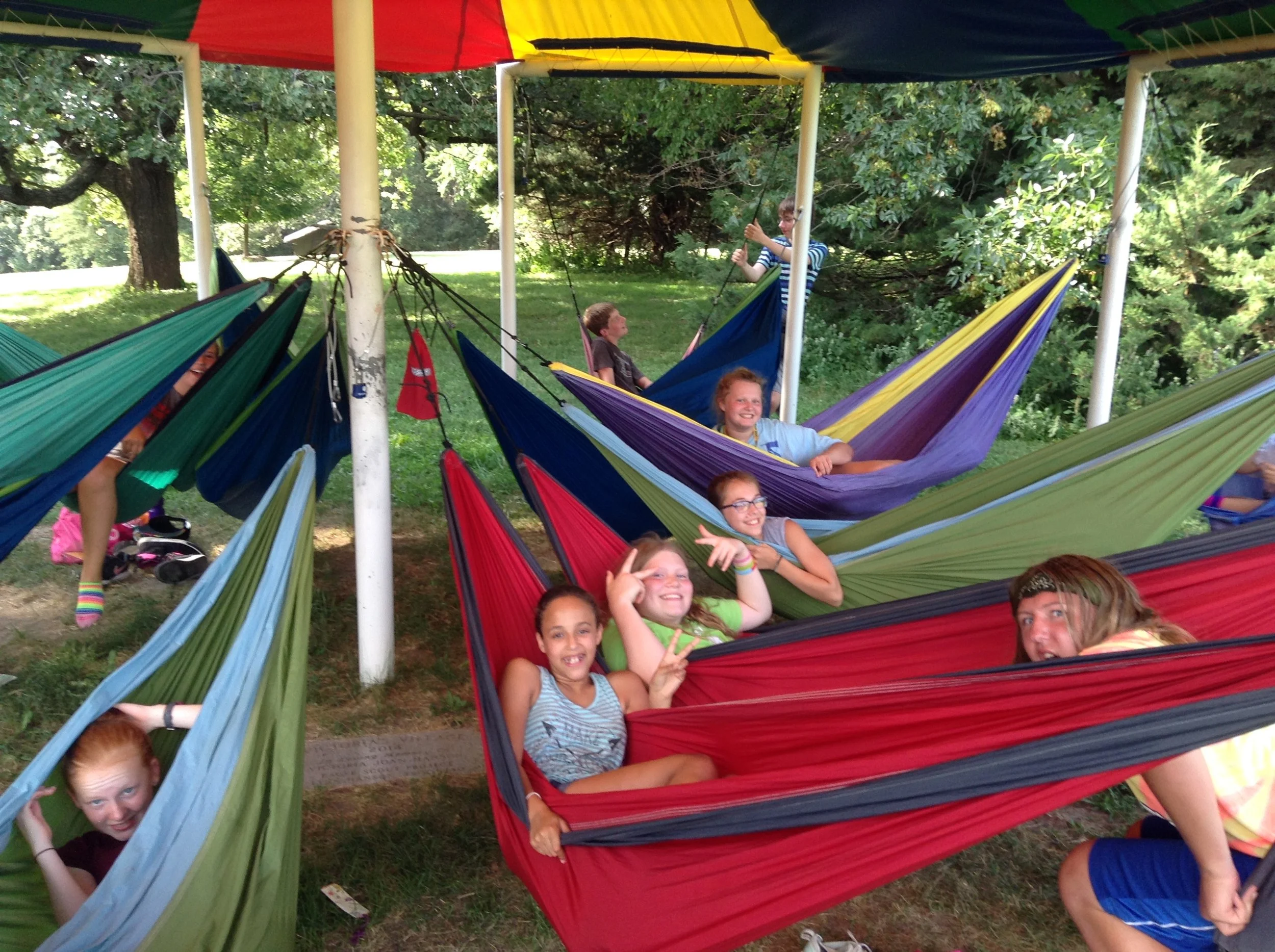 Children relaxing in colorful hammocks under a tent structure outdoors, surrounded by trees and green grass, enjoying a sunny day at a park or campsite.