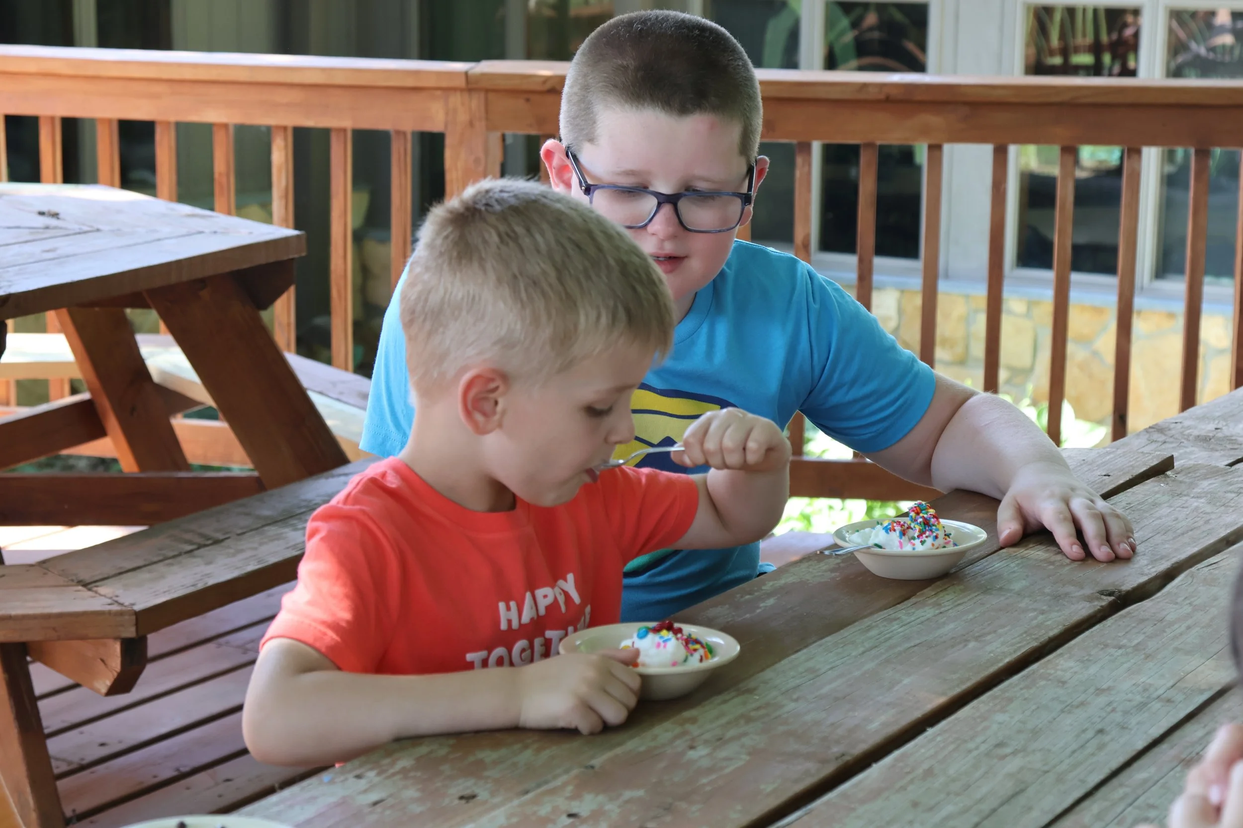 Two young boys, one older and one younger, enjoying ice cream with sprinkles at an outdoor wooden table on a porch.