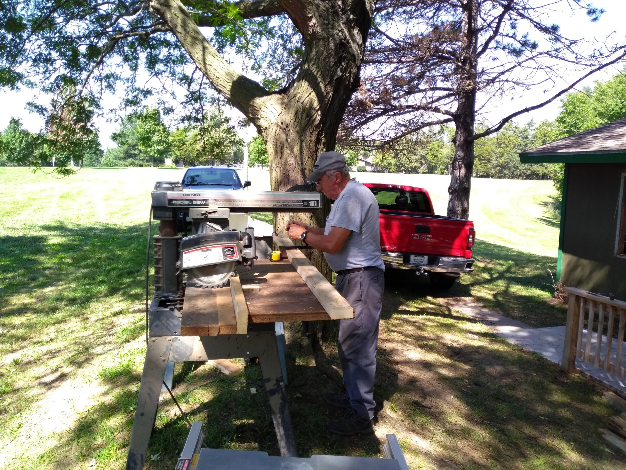 A man in a grey shirt and cap working with wood on a workbench outdoors. There is a large tree, a red truck, and a small green building in the background.