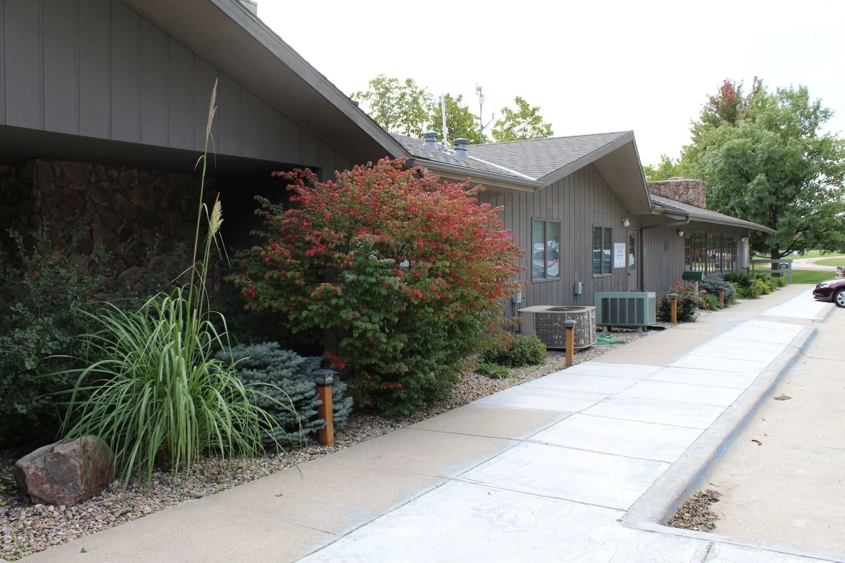 Exterior of a gray building with a sidewalk, landscaping, trees, and parked cars.