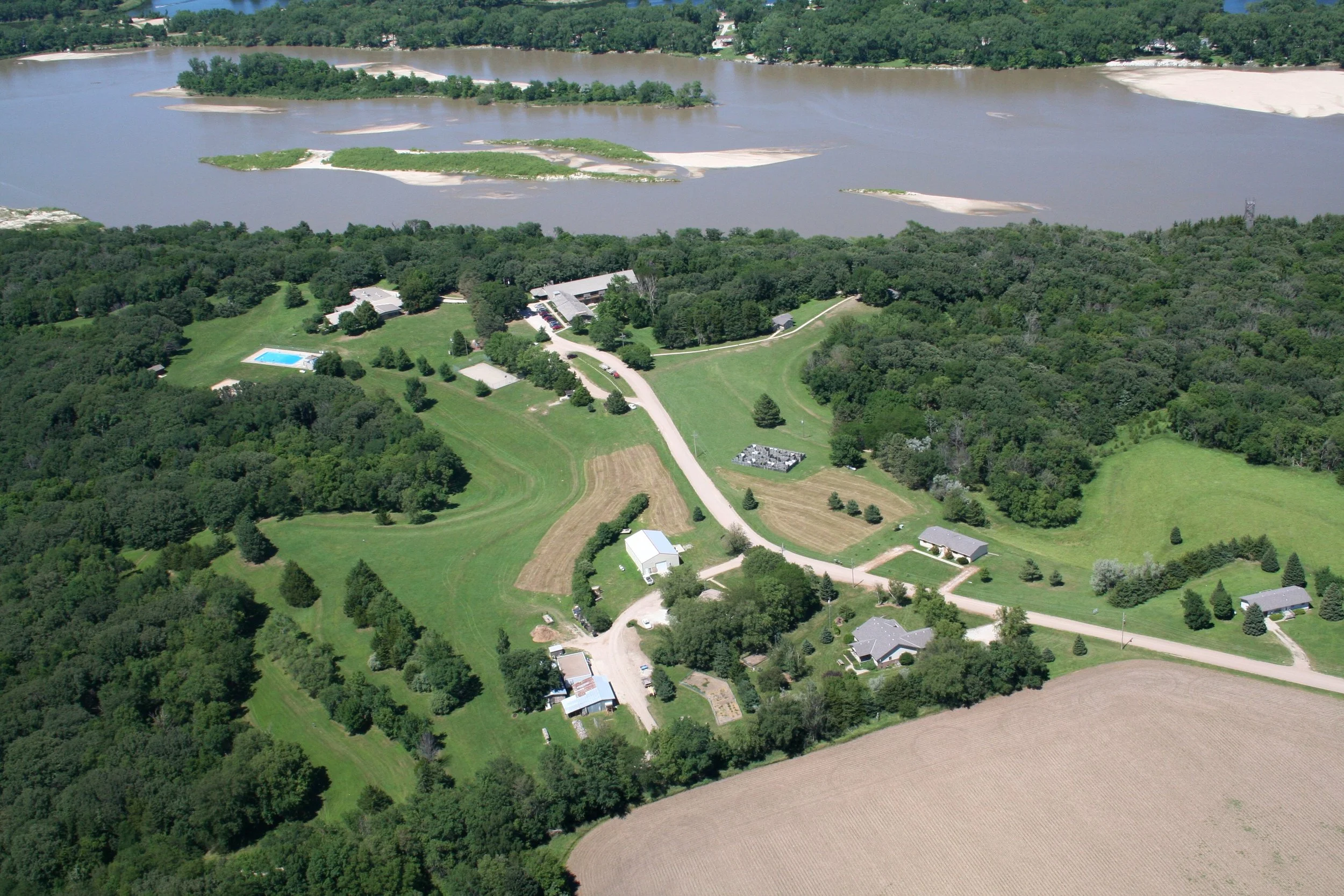 Aerial view of a tranquil natural space with cabins, conference center and other buildings as well as green fields, and a large river with islands.