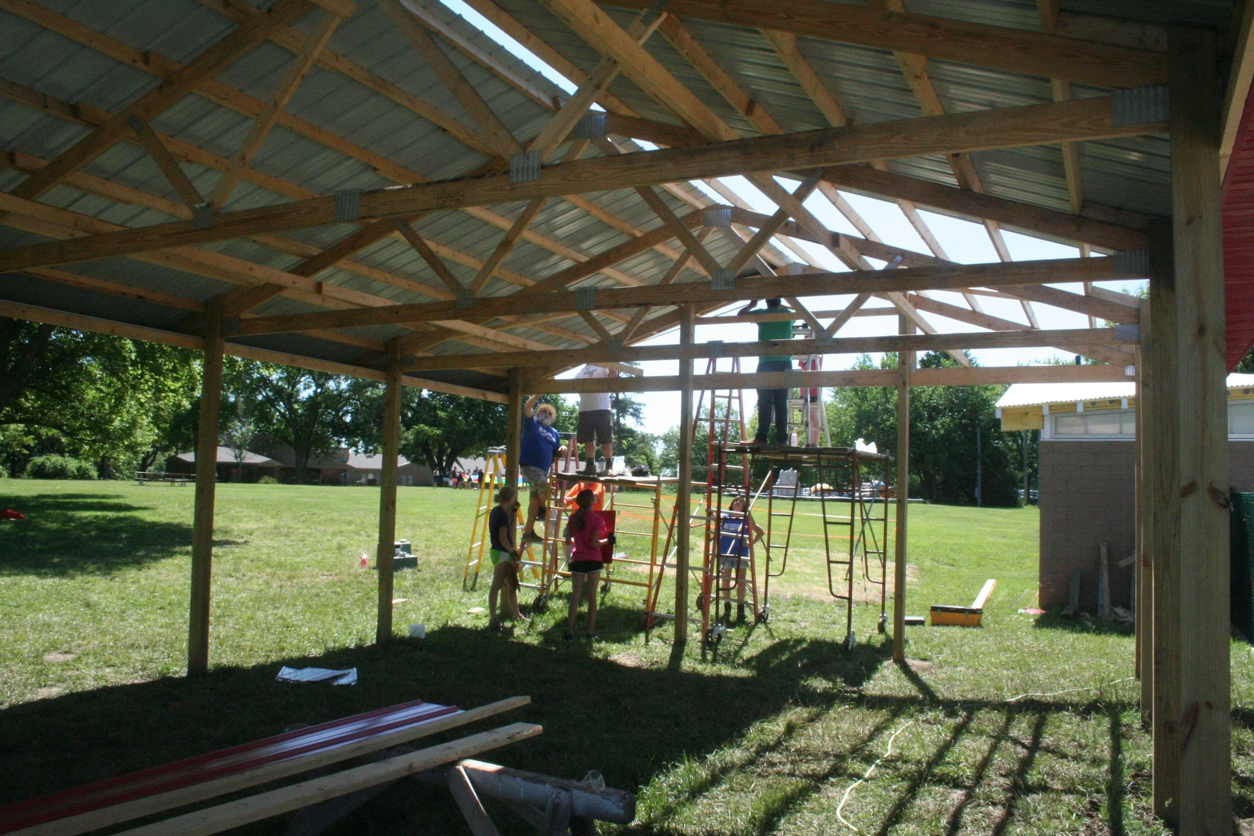 Kids and adults building a wooden pavilion in a park on a sunny day, with construction tools and materials around.