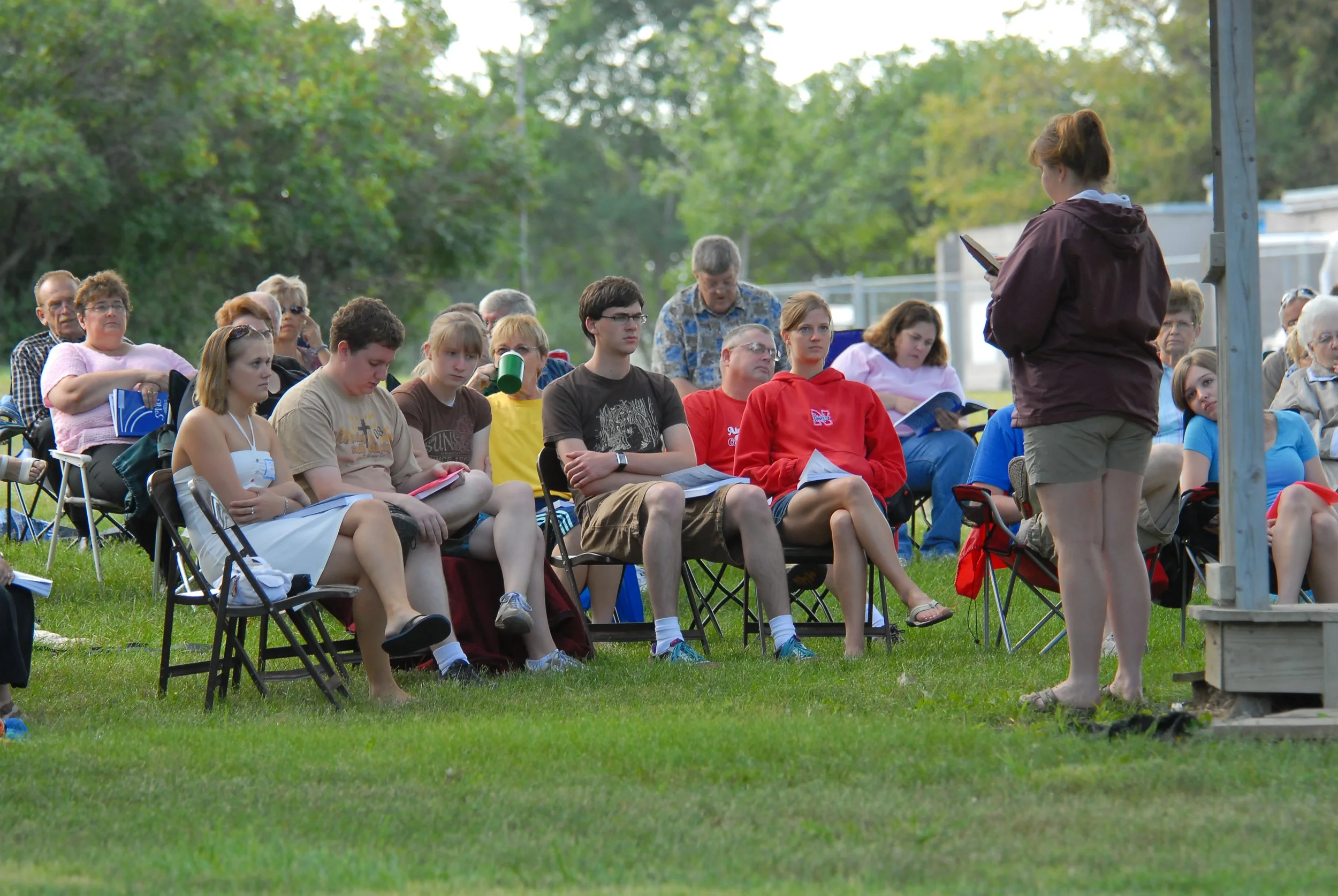 Group of people sitting on chairs outdoors, listening to a woman standing and reading from a book or paper, with trees and grass in the background.