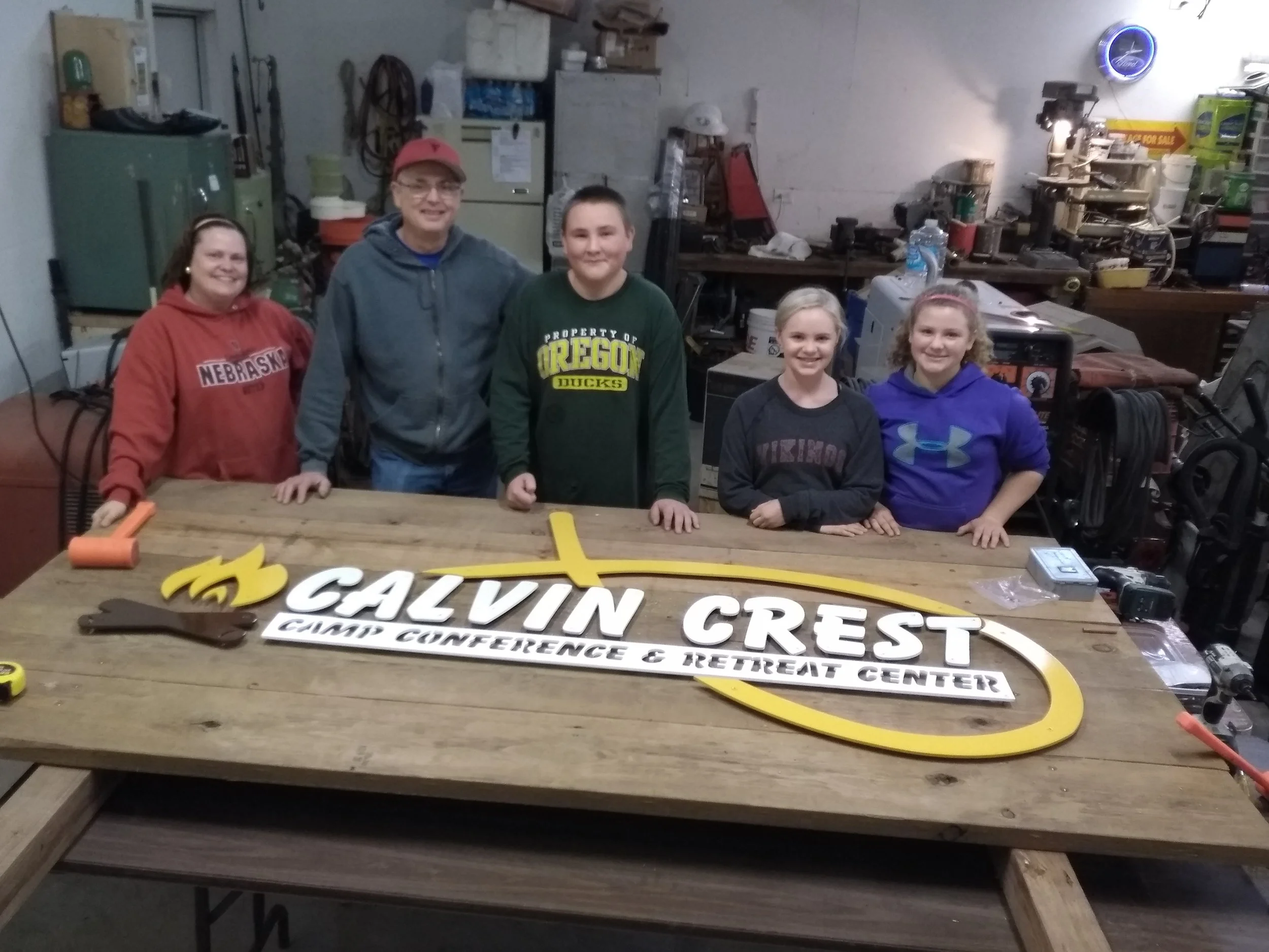 Group of six people standing behind a large wooden table with a decorated sign that reads "Calvin Crest Camp Conference & Retreat Center" in a workshop or garage setting.