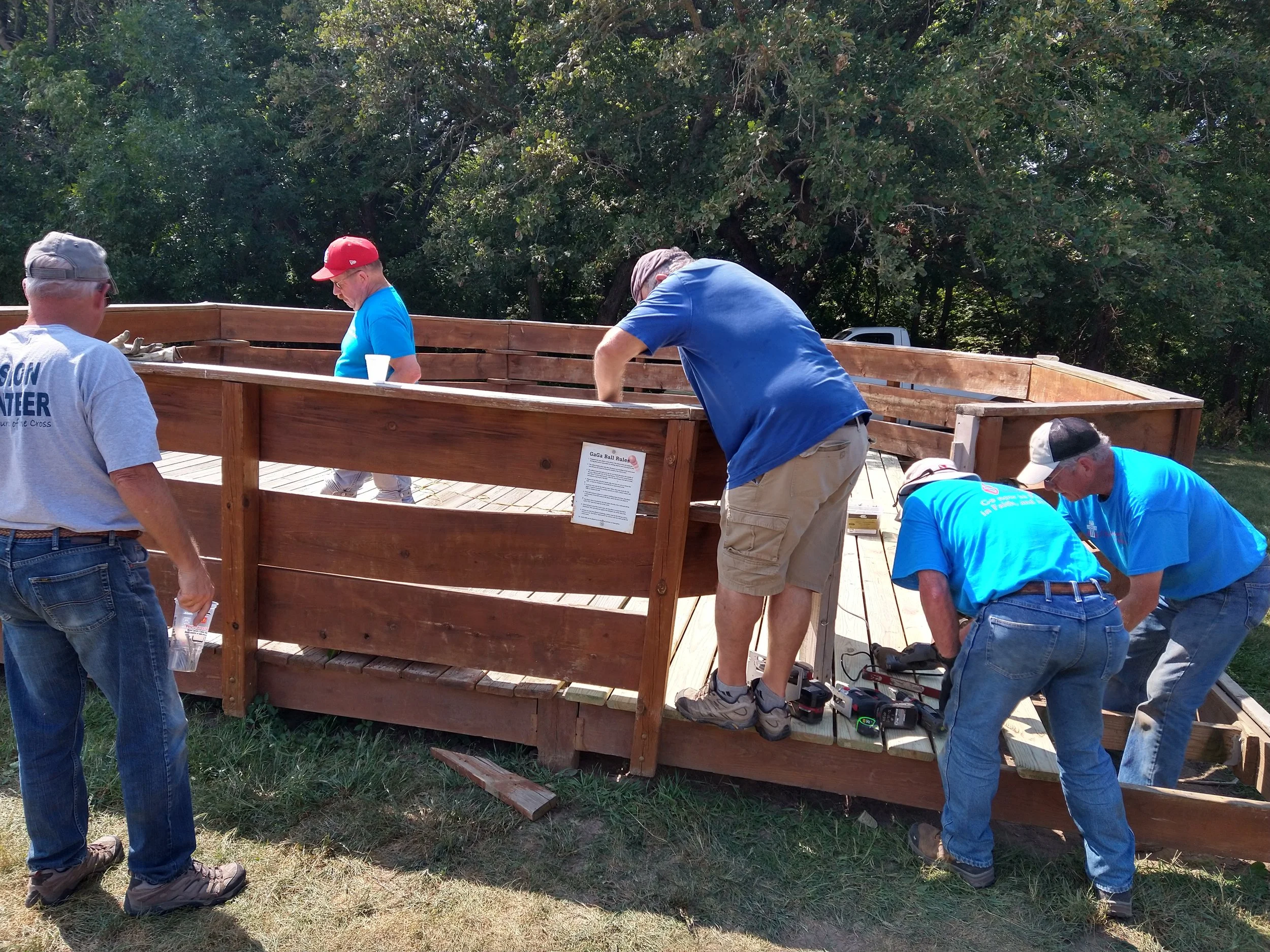 Group of people working on building a wooden platform outdoors with tools and instructions, surrounded by trees.