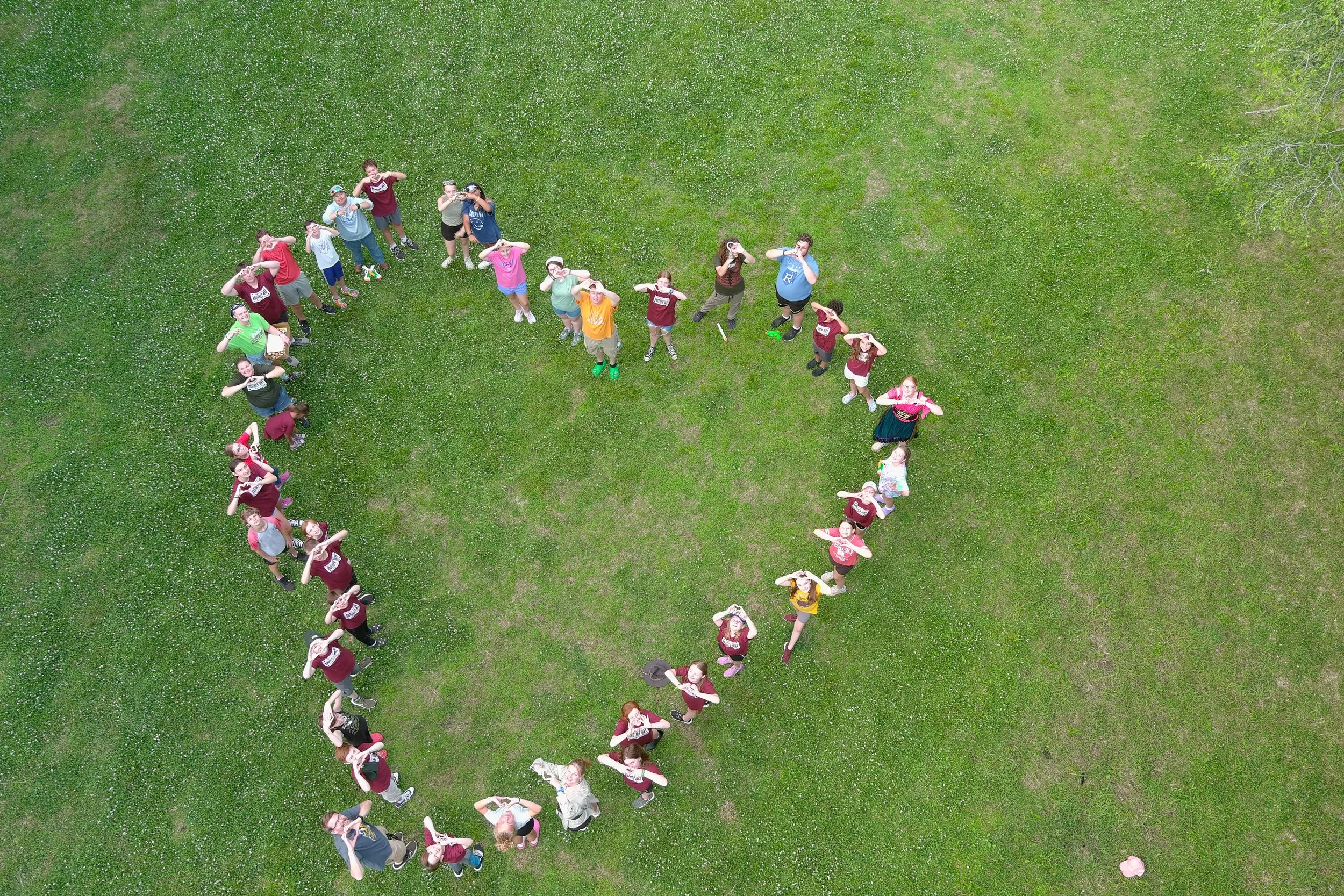 Group of people forming the shape of a heart on a grassy field, looking up at the camera.