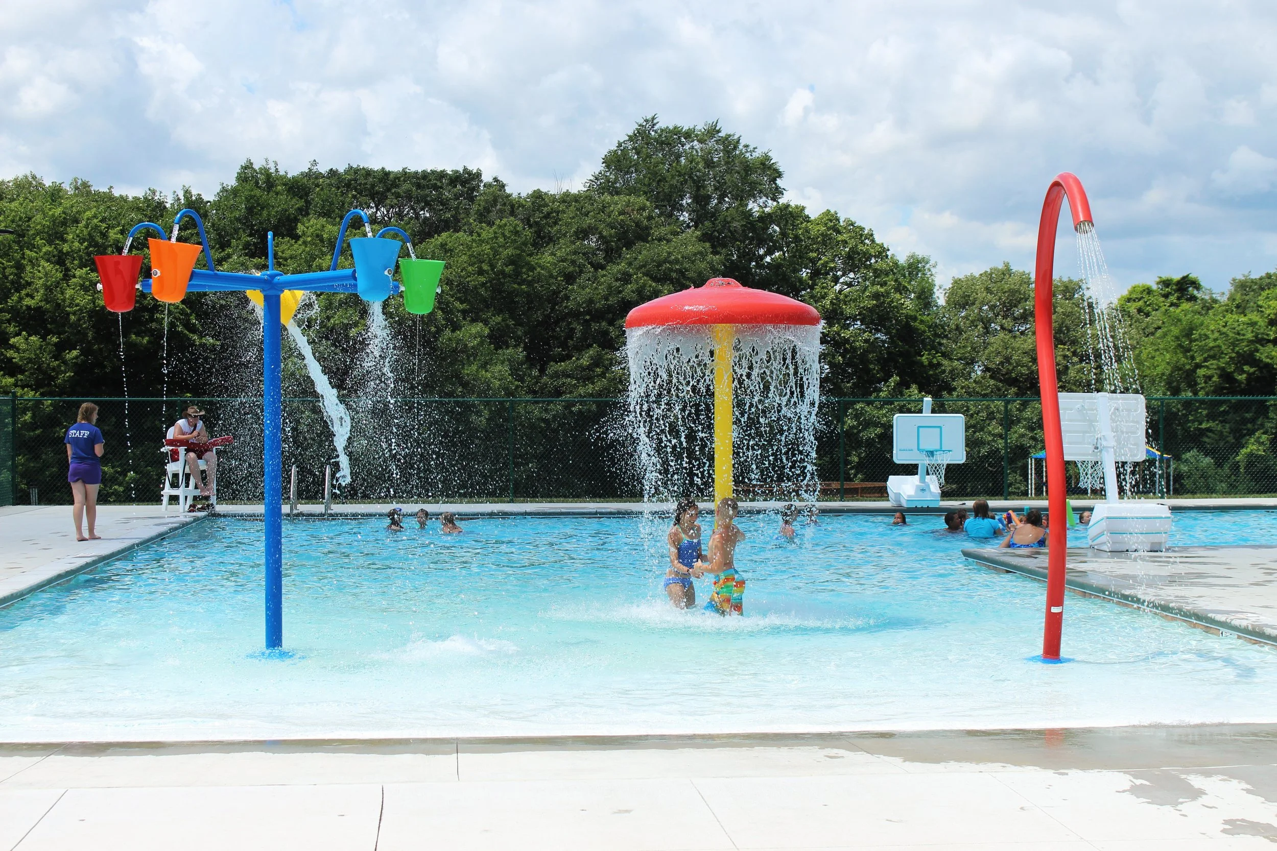 Children playing and splashing in an outdoor water park pool with colorful water features and a basketball hoop, surrounded by green trees.