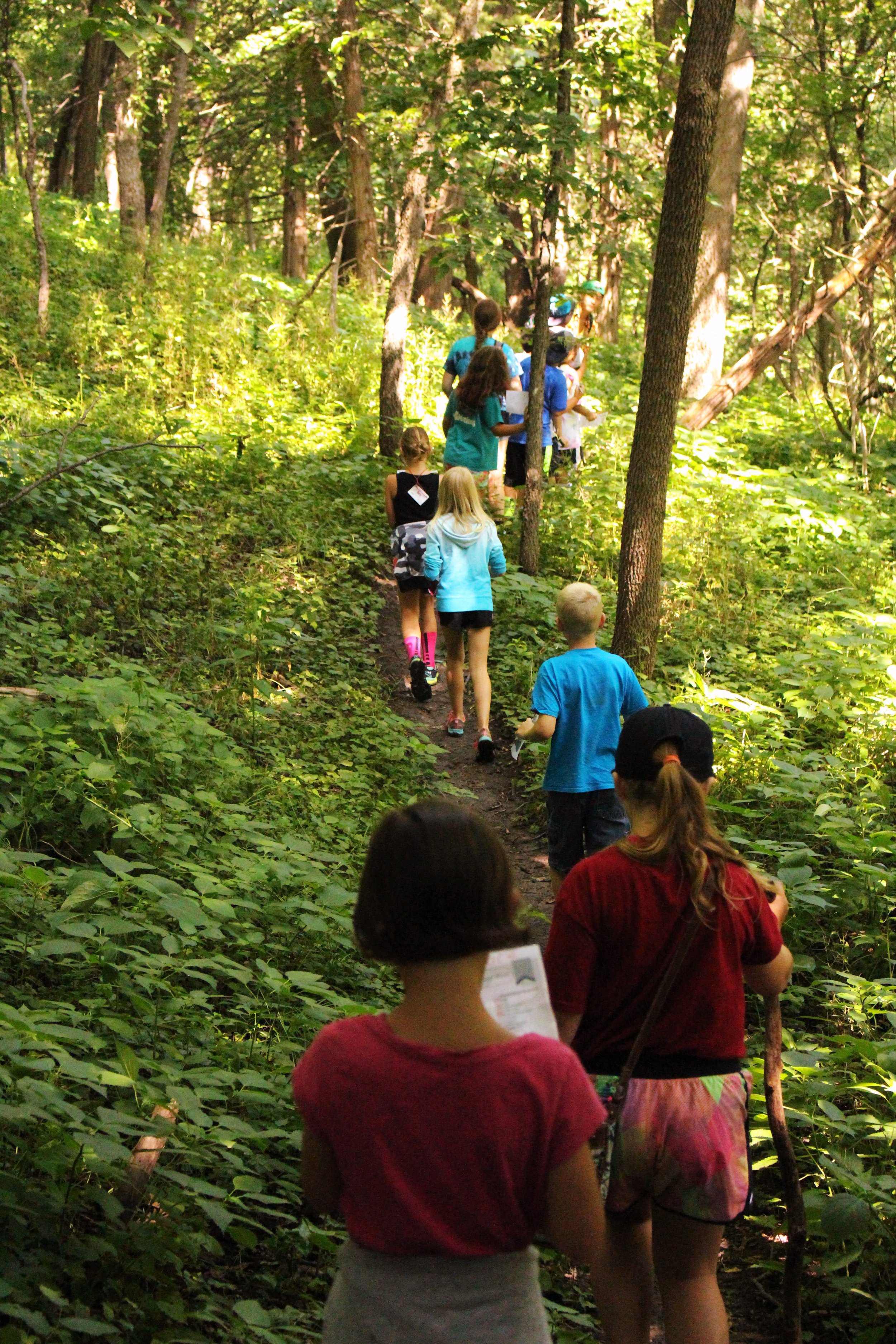 A group of children and adults walking in a single file line on a narrow trail through a dense green forest.