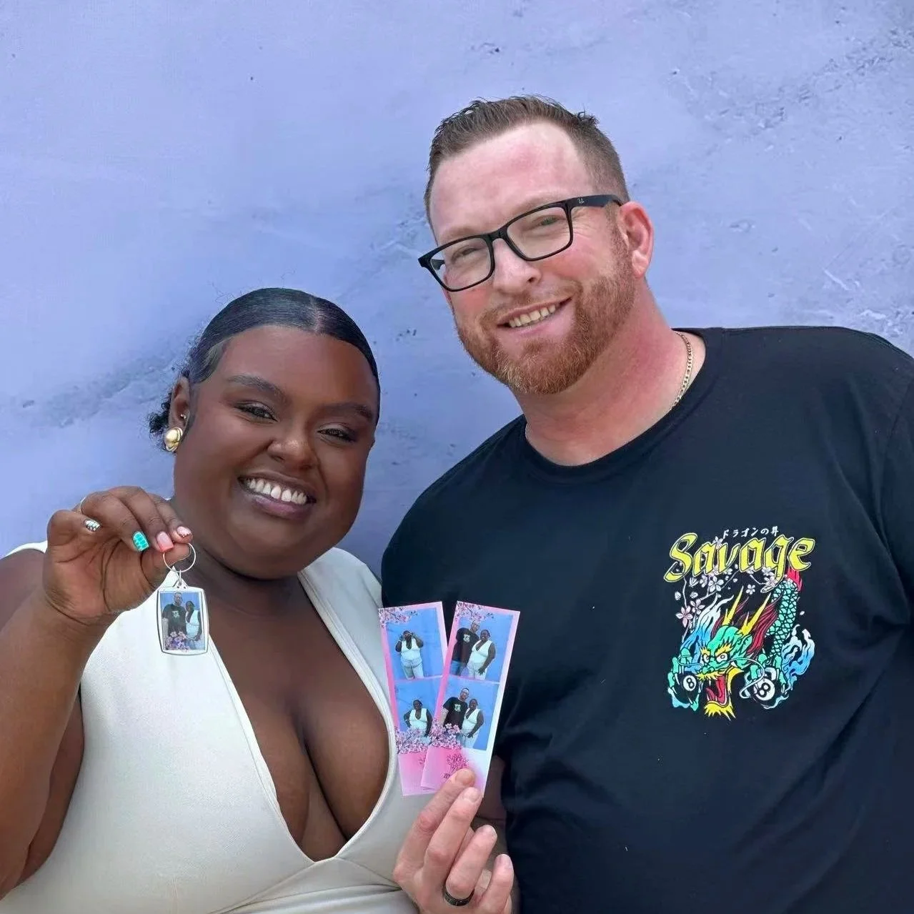 A smiling woman with dark hair holds a keychain with a photo, standing next to a smiling man wearing glasses with a graphic T-shirt, both holding photo booth pictures, standing against a purple wall.