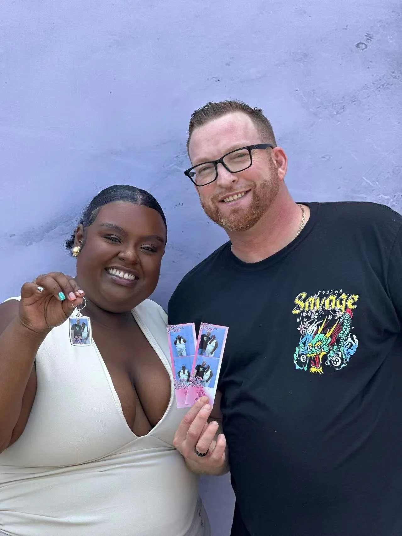 A woman and a man stand together smiling, holding photo strips and a keychain with photo strips. The woman is wearing a cream-colored top with a deep neckline, earrings, and has her hair slicked back. They are standing against a purple wall.