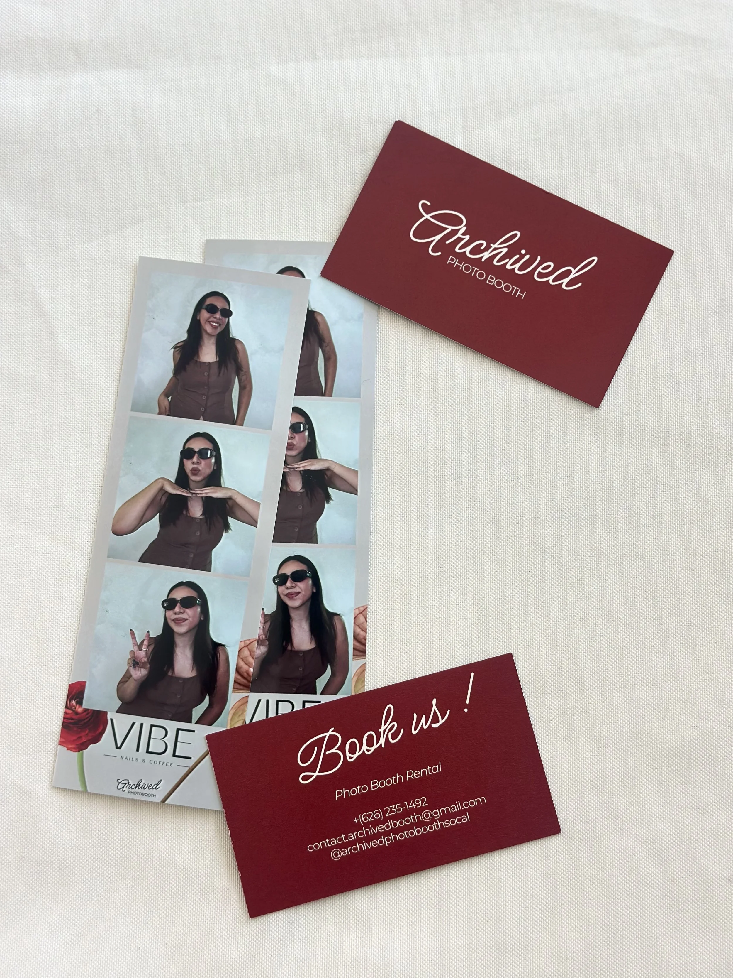 Photo booth strip with four images of a woman wearing sunglasses and a brown top, making different poses. Two red business cards, one reading 'Archived Photo Booth' and the other with booking information, are placed on a white surface.