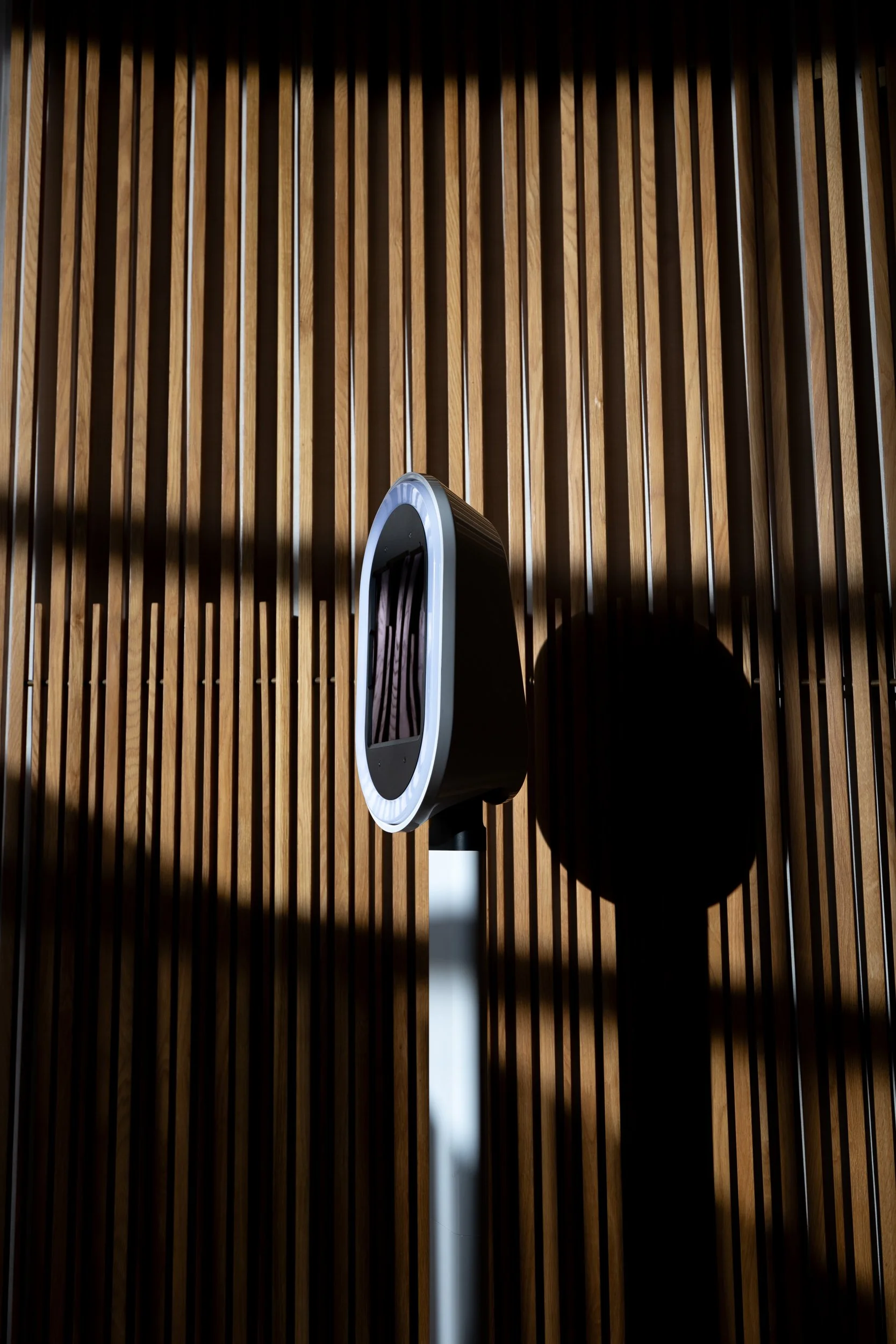 A black and silver electric vehicle charger mounted on a white stand against a vertical wooden slat wall, casting a shadow.