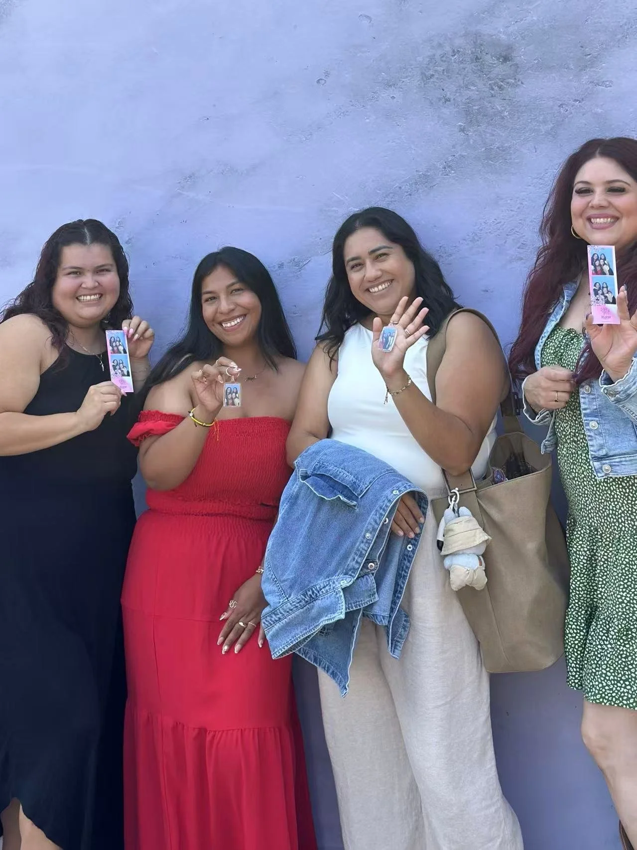 Four women smiling and holding photo booth strips, standing against a blue wall, dressed casually and carrying bags.
