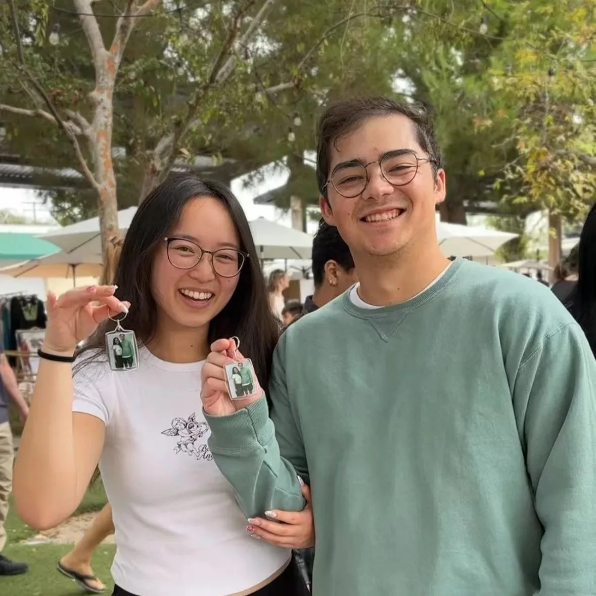 A smiling woman with dark hair holds a keychain with a photo, standing next to a smiling man wearing glasses with a graphic T-shirt, both holding photo booth pictures, standing against a purple wall.