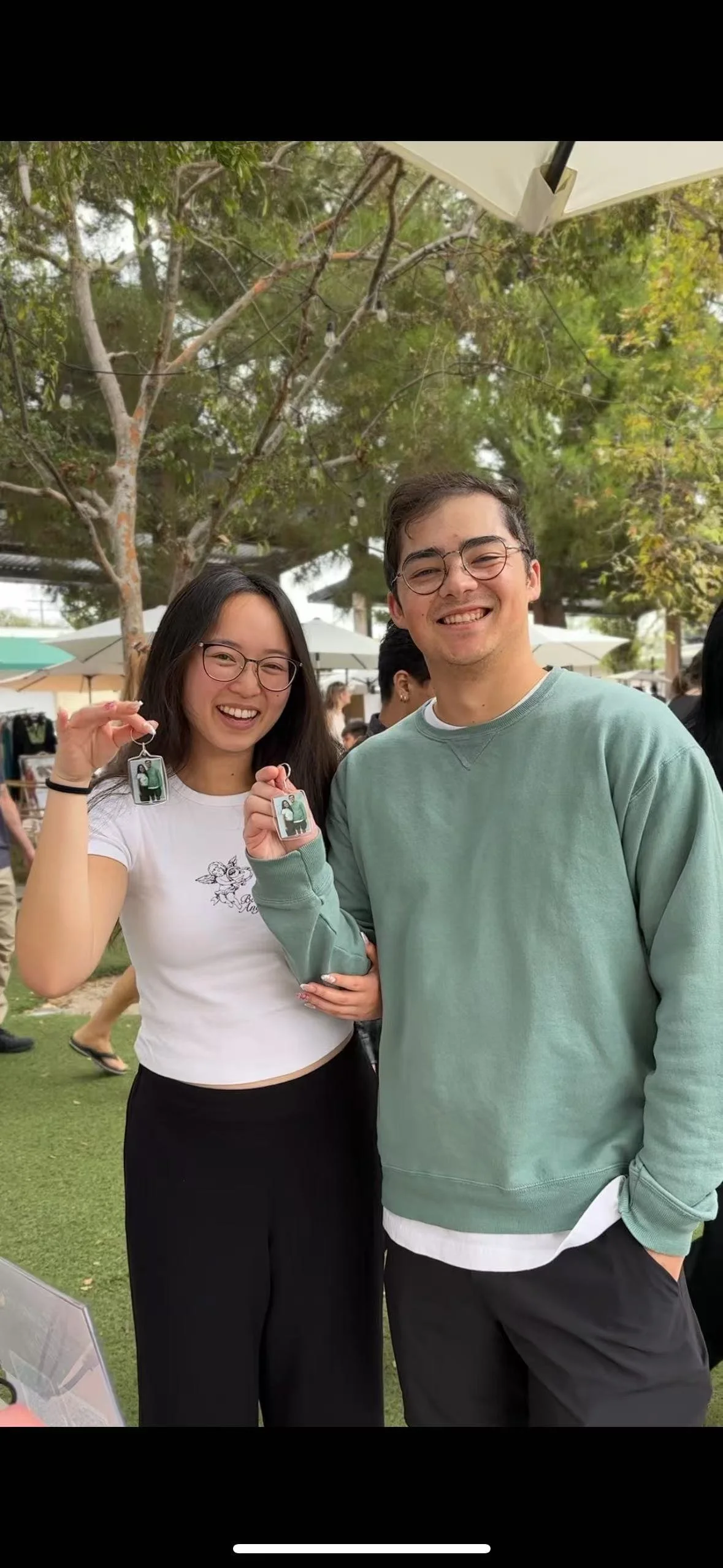 Two young adults smiling and holding up keychains with photos of themselves outdoors at a gathering with trees and umbrellas in the background.