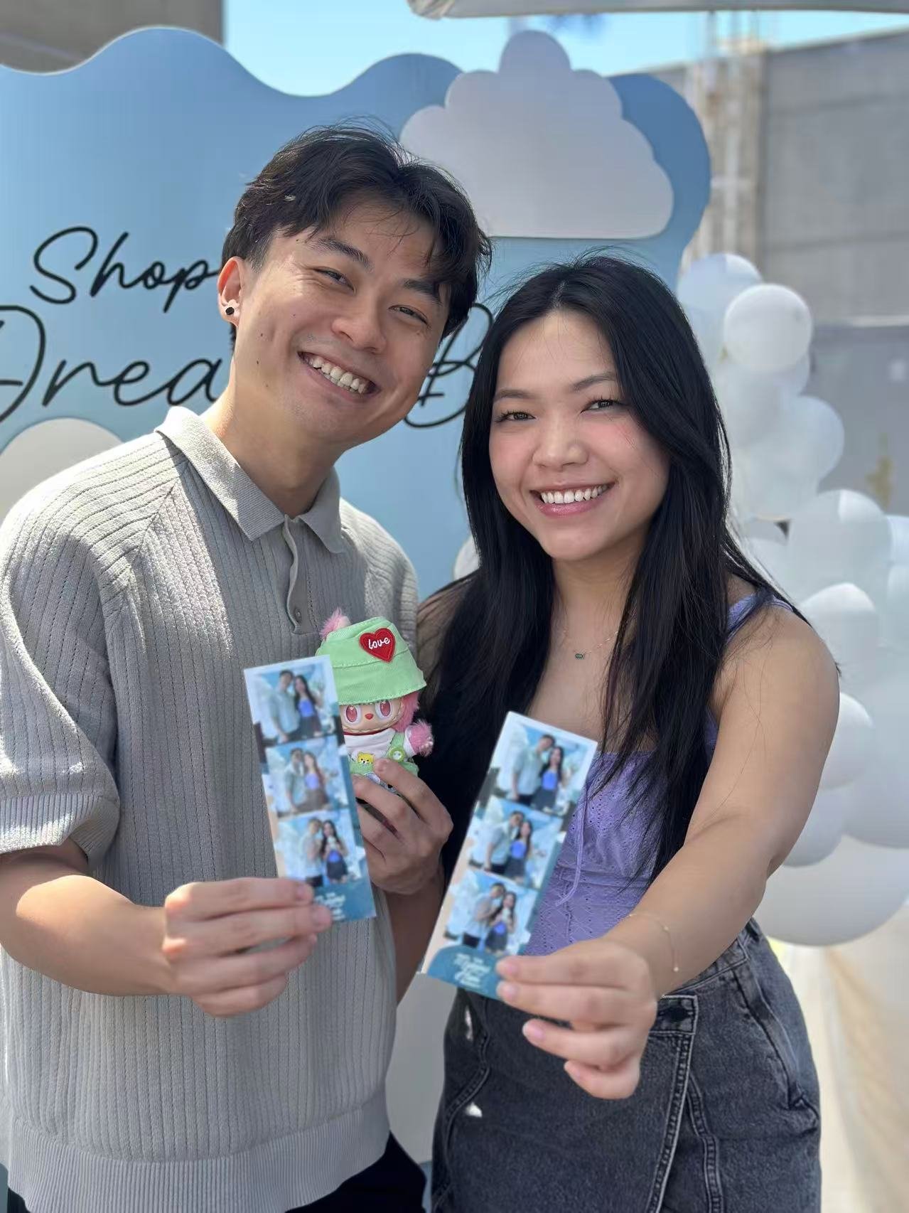A smiling young couple holding photo booth strips and a plush toy at a dream-themed event with blue and white cloud decorations.