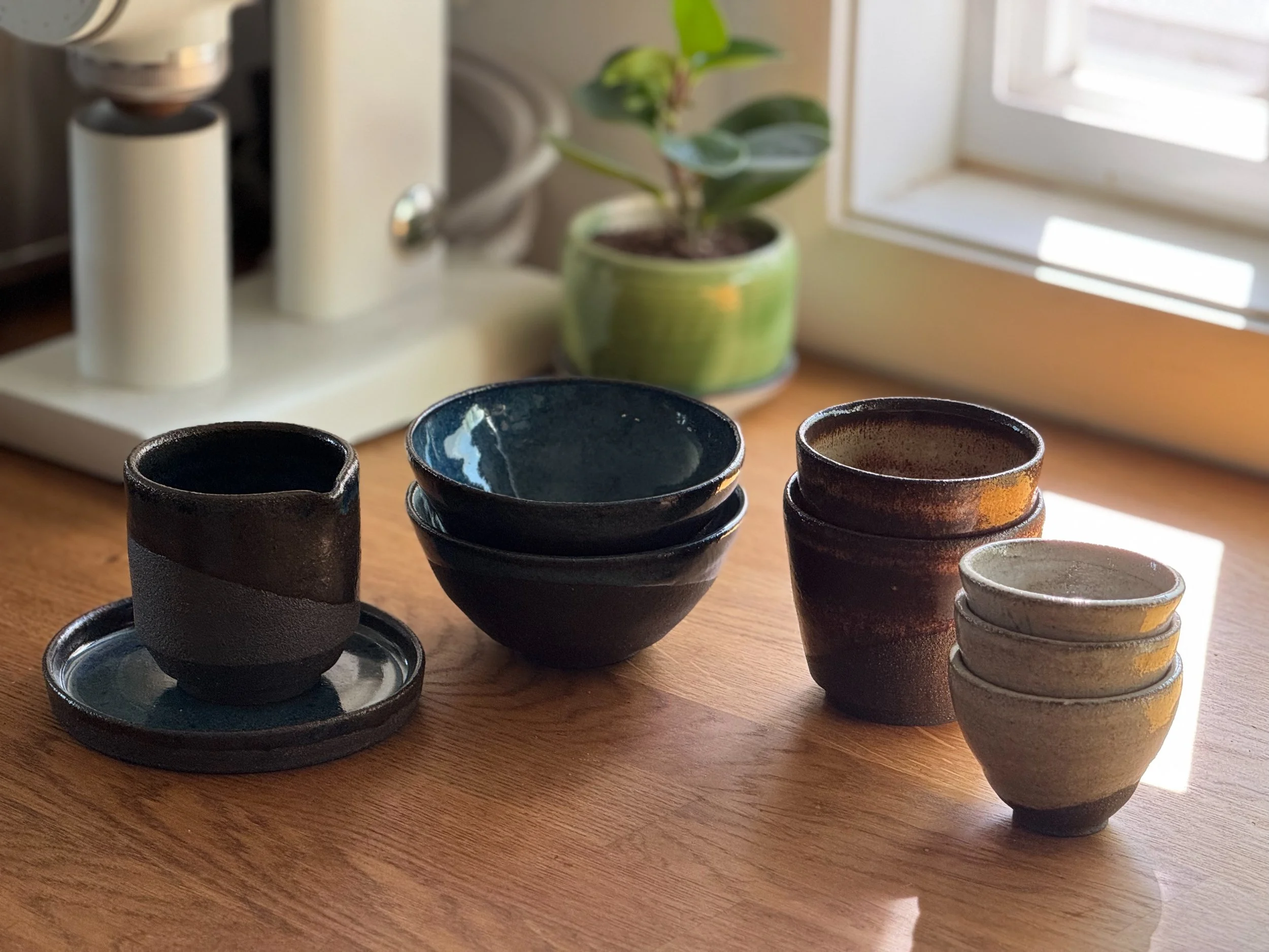 Stacked cups of various colors on a wooden table in front of a window with blinds, with a potted plant and candles in the background.
