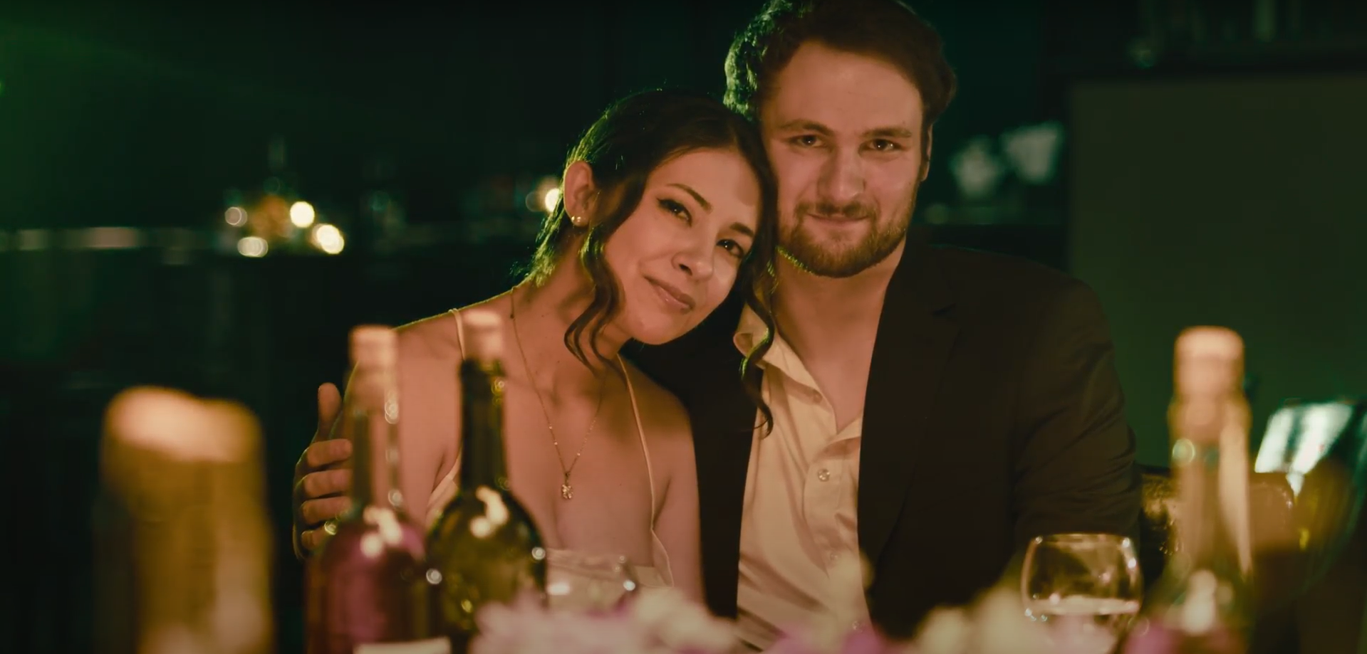 A couple sitting at a dinner table, smiling and cuddling, surrounded by lit candles and bottles of wine or beer.