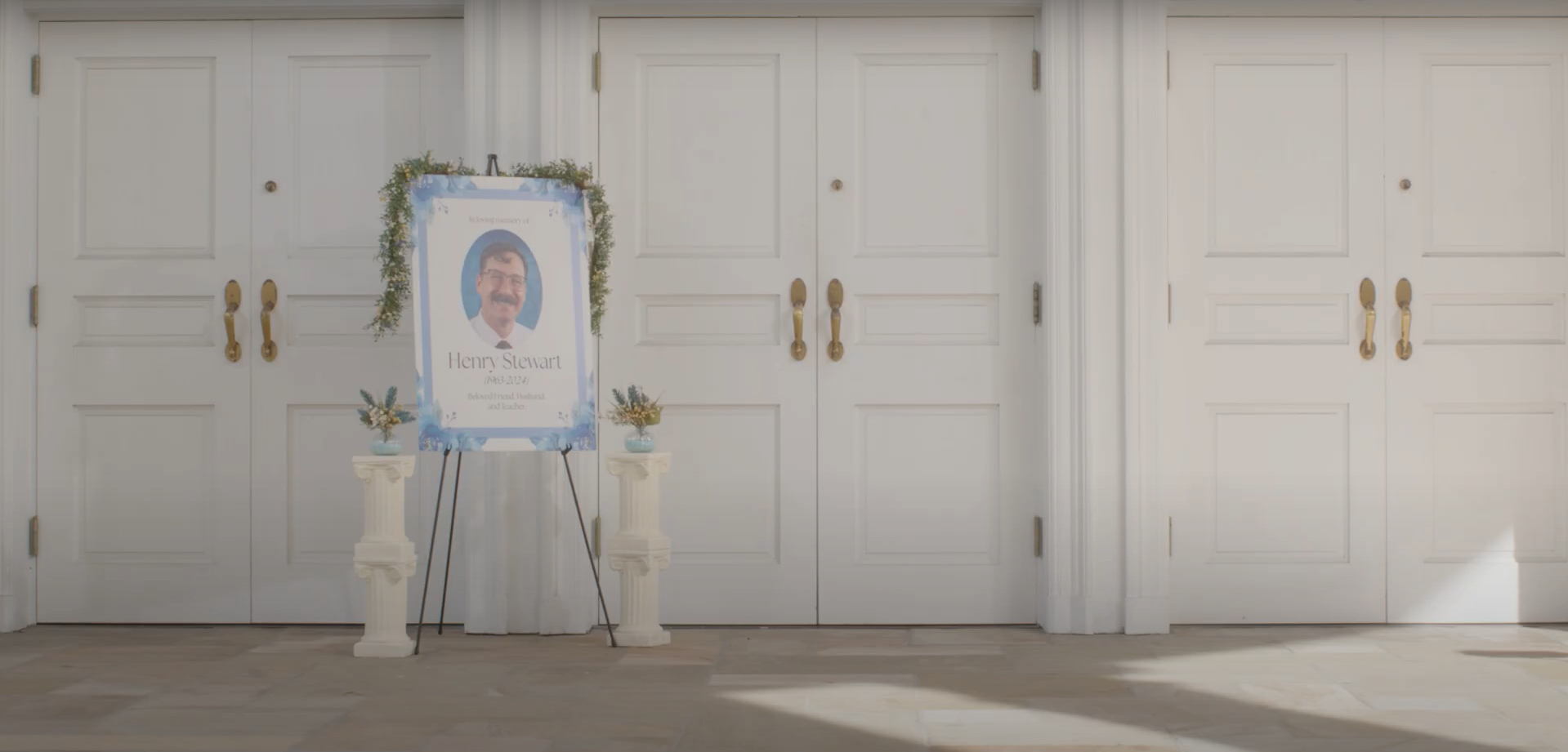 Funeral or memorial setup with a picture of Henry Stewart on a stand, flanked by small flower arrangements on white pedestals, against white double doors with brass handles.