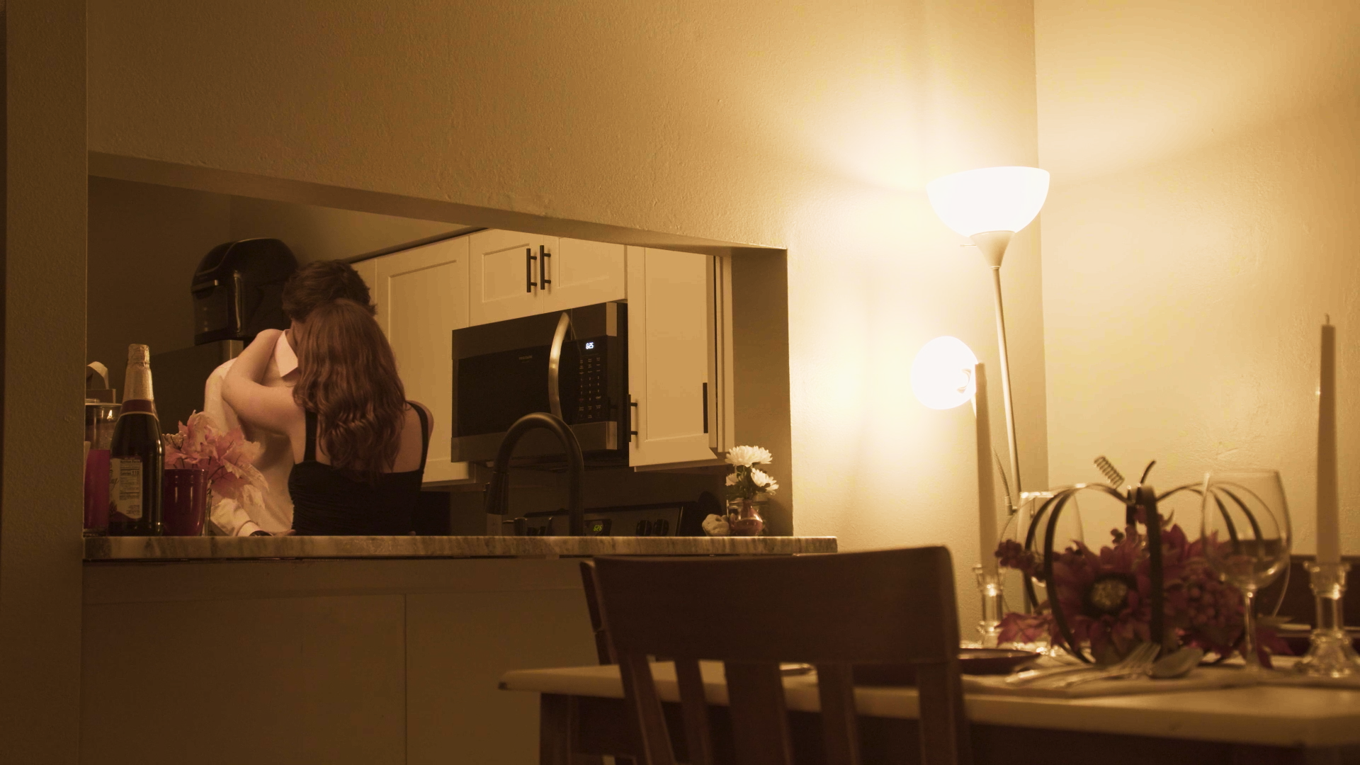 Two people sharing an intimate kiss in a kitchen, with warm lighting, a microwave, flowers, and a bottle of champagne on the counter, as seen through an open living room doorway.