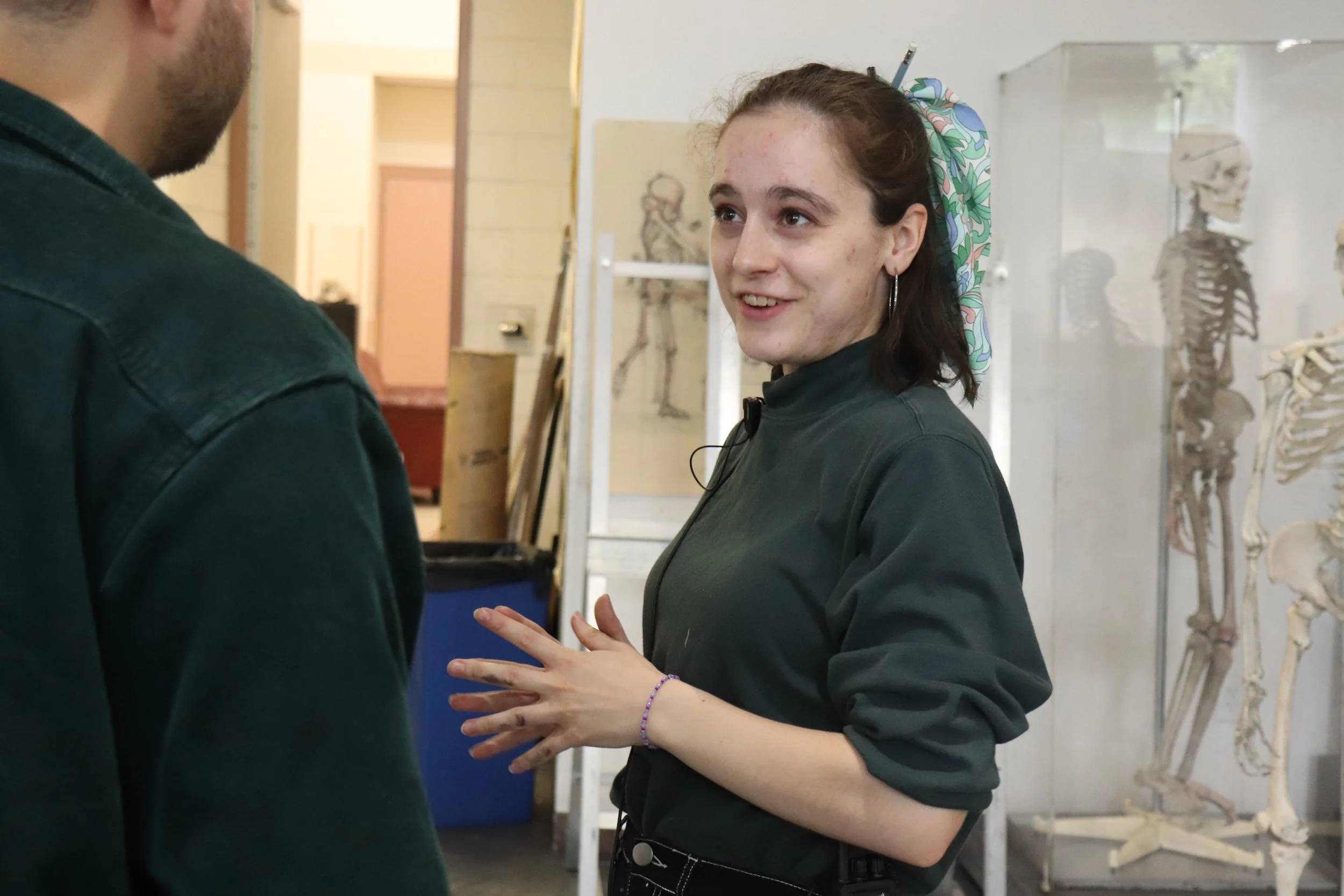 A young woman with brown hair, wearing a dark green jacket and a patterned headscarf, is talking to a man whose face is not visible. She appears to be explaining something with her hands in a museum or educational setting with skeletons and anatomy drawings in the background.