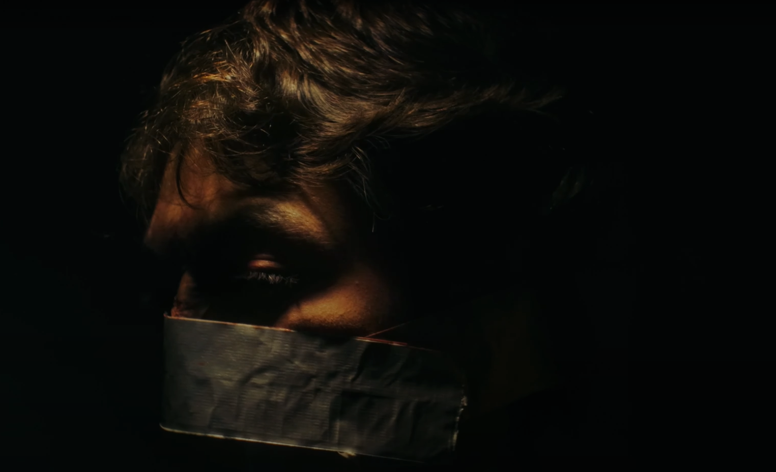 A person with wavy hair wearing a metallic face mask, illuminated against a dark background.