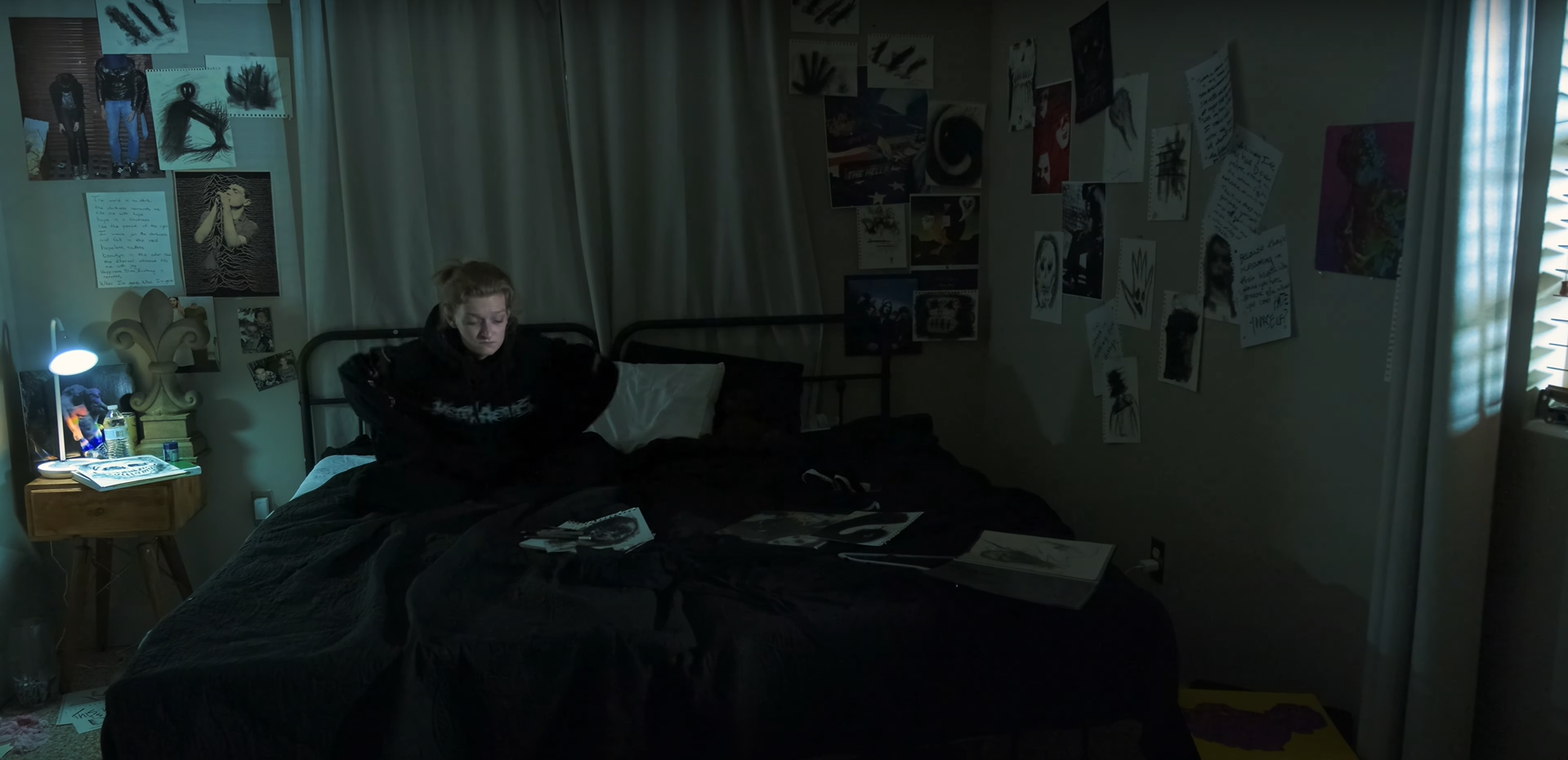 A young woman sits on a bed in a dimly lit bedroom, surrounded by artwork and photographs on the walls, with a bedside table holding a lamp and water bottles.