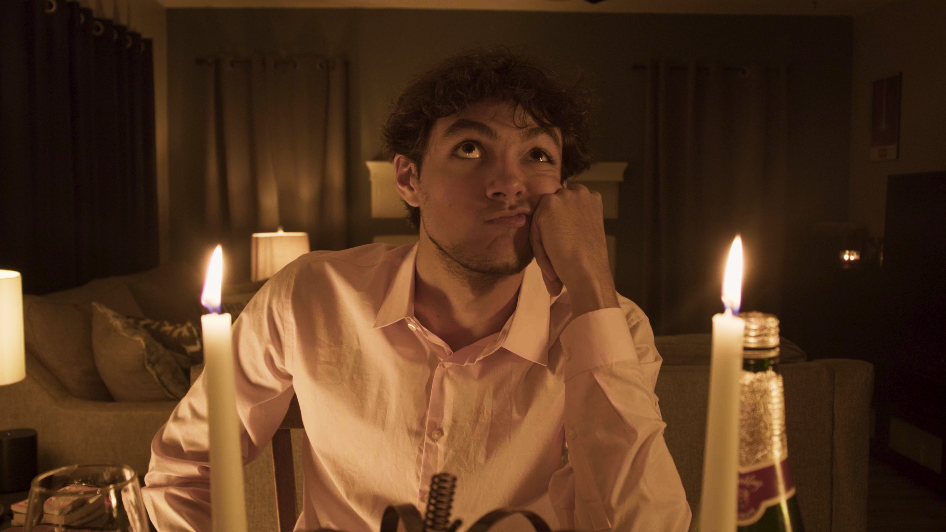 A young man sitting at a dining table with a neutral expression, resting his chin on his hand, in a dimly-lit room illuminated by candlelight.